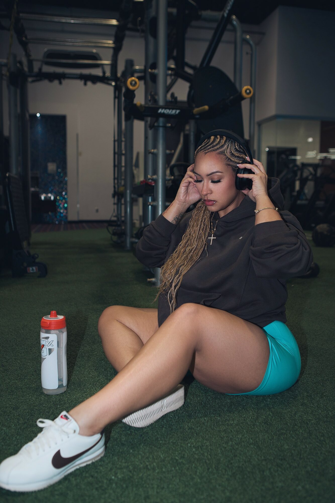 Young woman sitting on gym floor, adjusting her hair, with water bottle nearby, gym equipment in background.