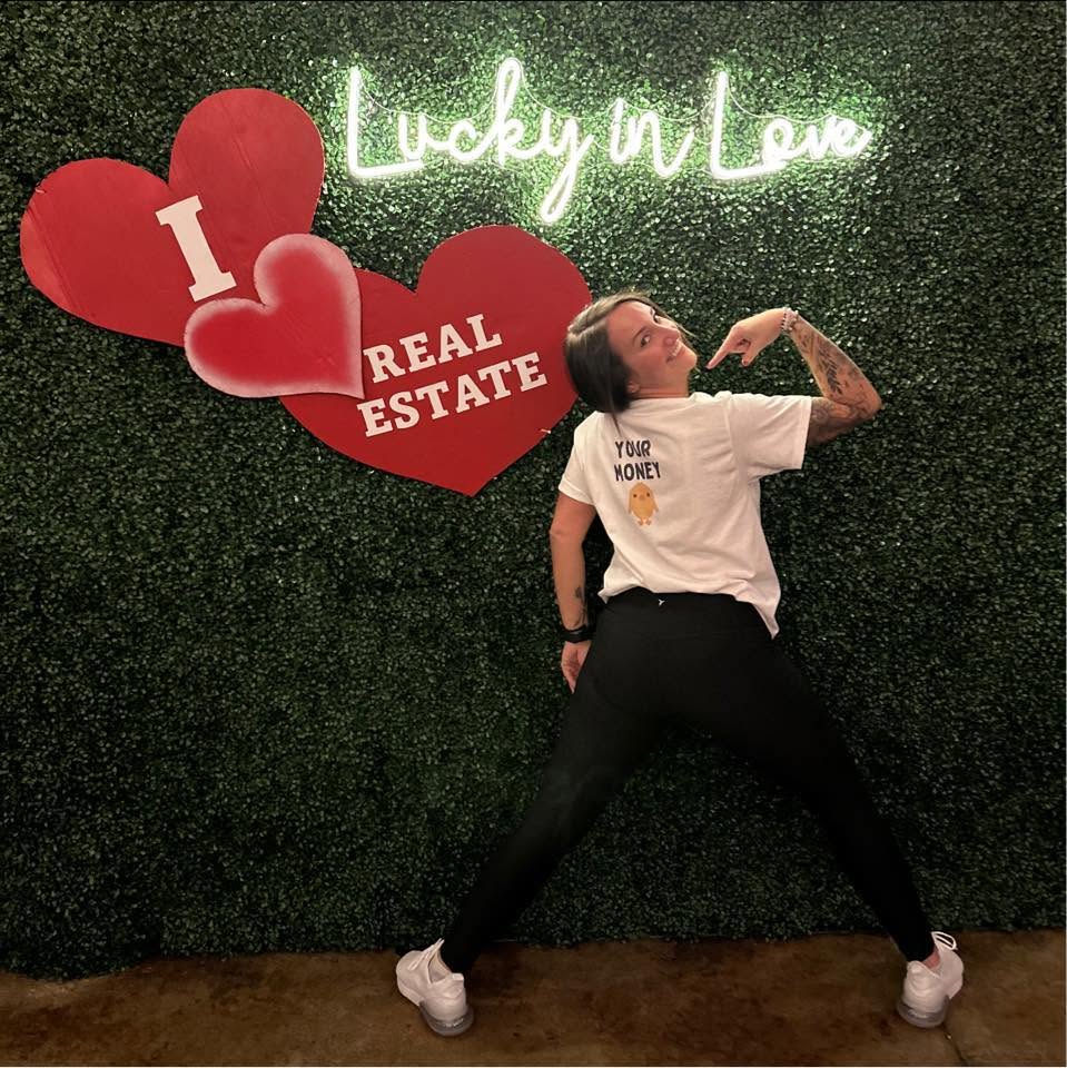 Person with tattoos poses with legs apart in front of a green wall with neon sign and heart-shaped signs, holding a pose.