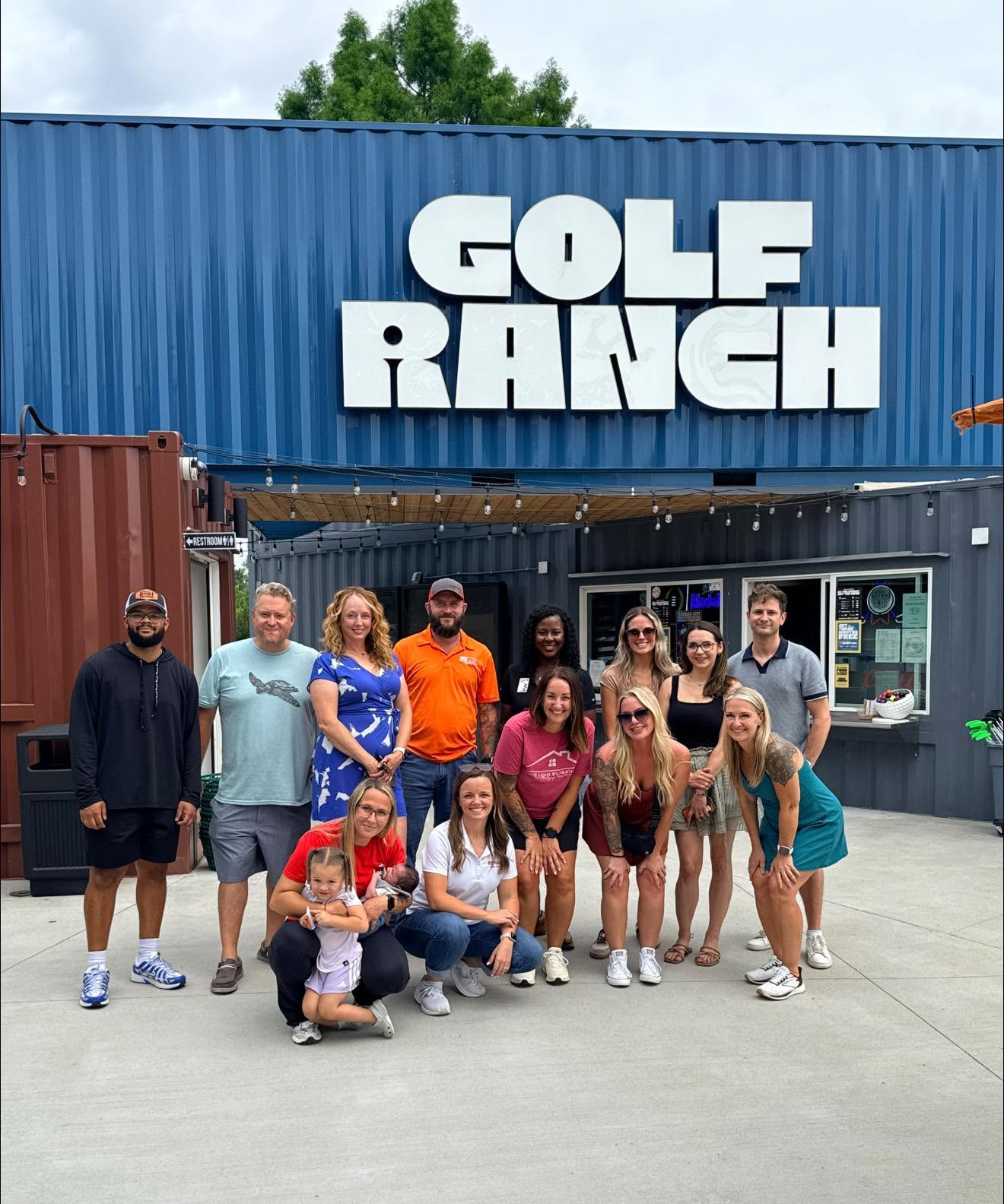 Group of people standing outside GOLF RANCH building, some smiling, some with hands on hips, in front of blue and black walls.