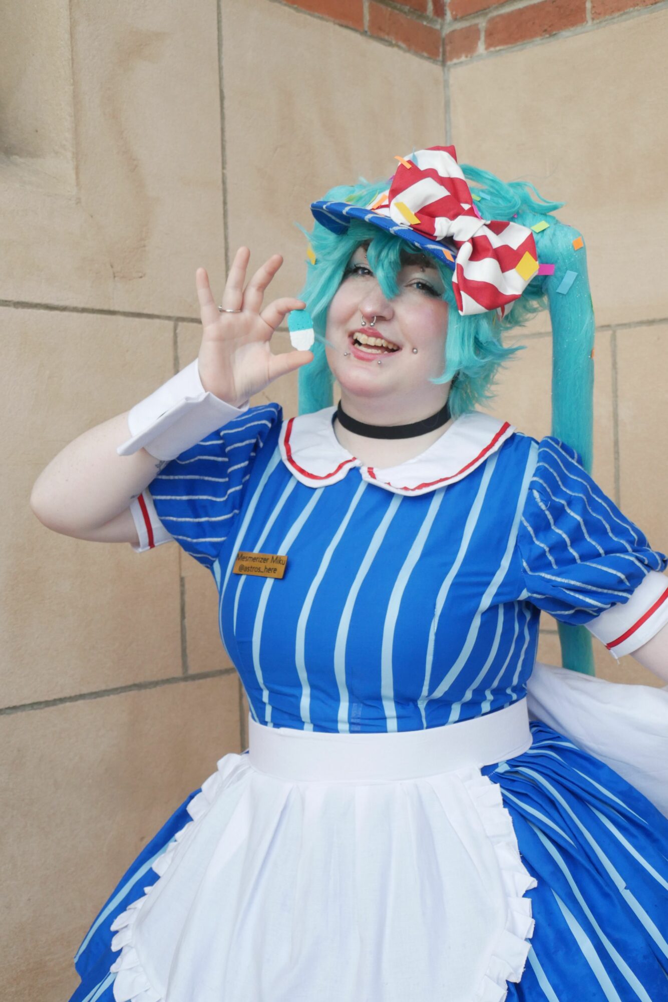 Person dressed in a colorful costume with a striped blue and white top, white skirt, and a multicolored wig, smiling and making a peace sign.