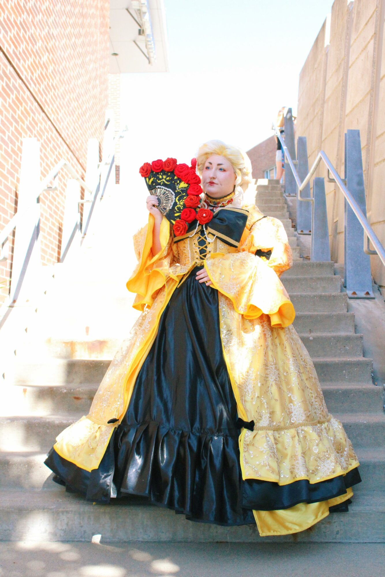 Person in elaborate yellow and black gown holding a bouquet of red roses, standing on outdoor stairs.