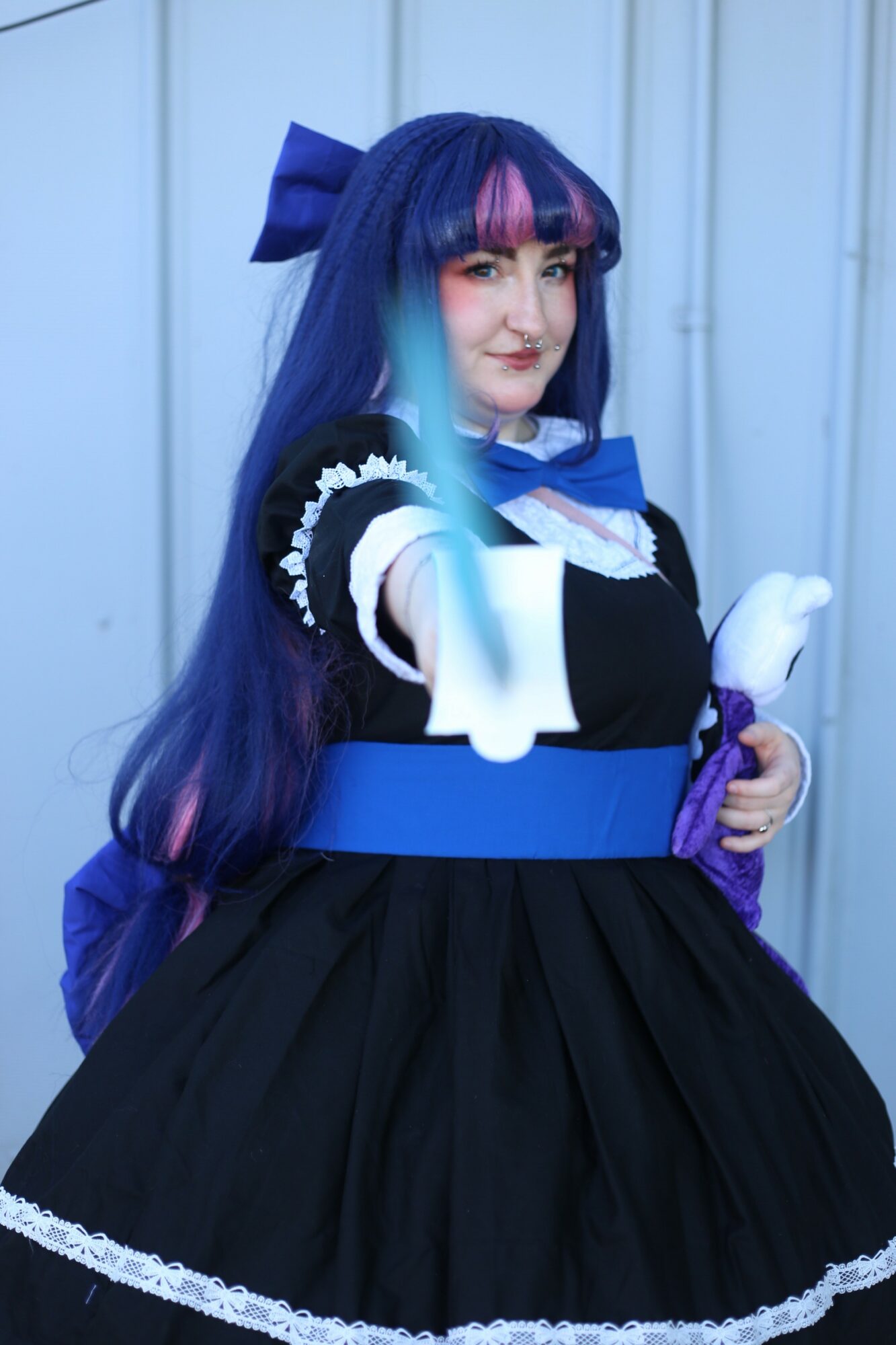 Person in costume with long blue and purple hair, wearing a black dress with blue and white accents, holding a paper fan.