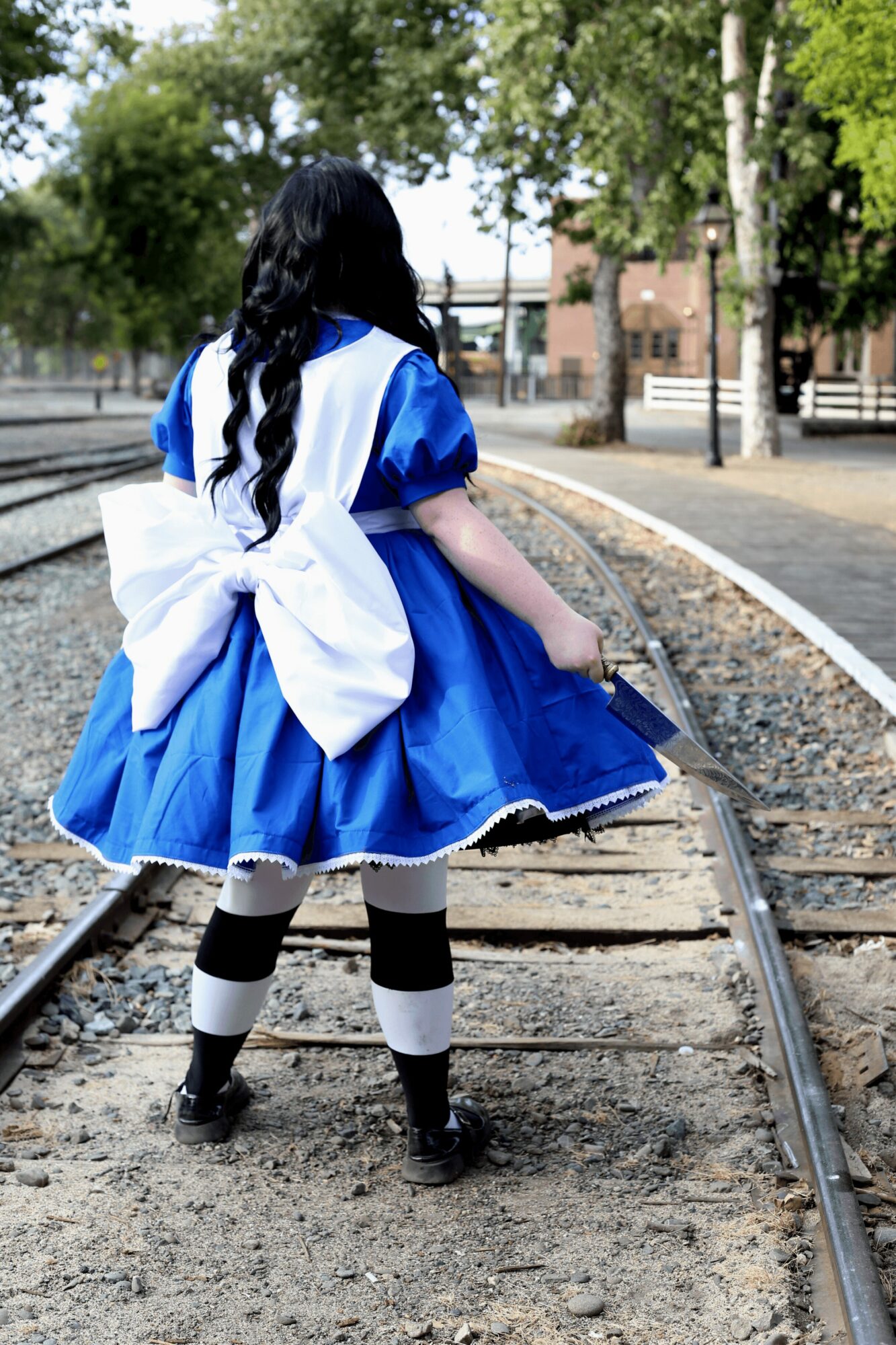 Girl in blue dress with large white bow standing on train tracks outdoors, trees and buildings in background.