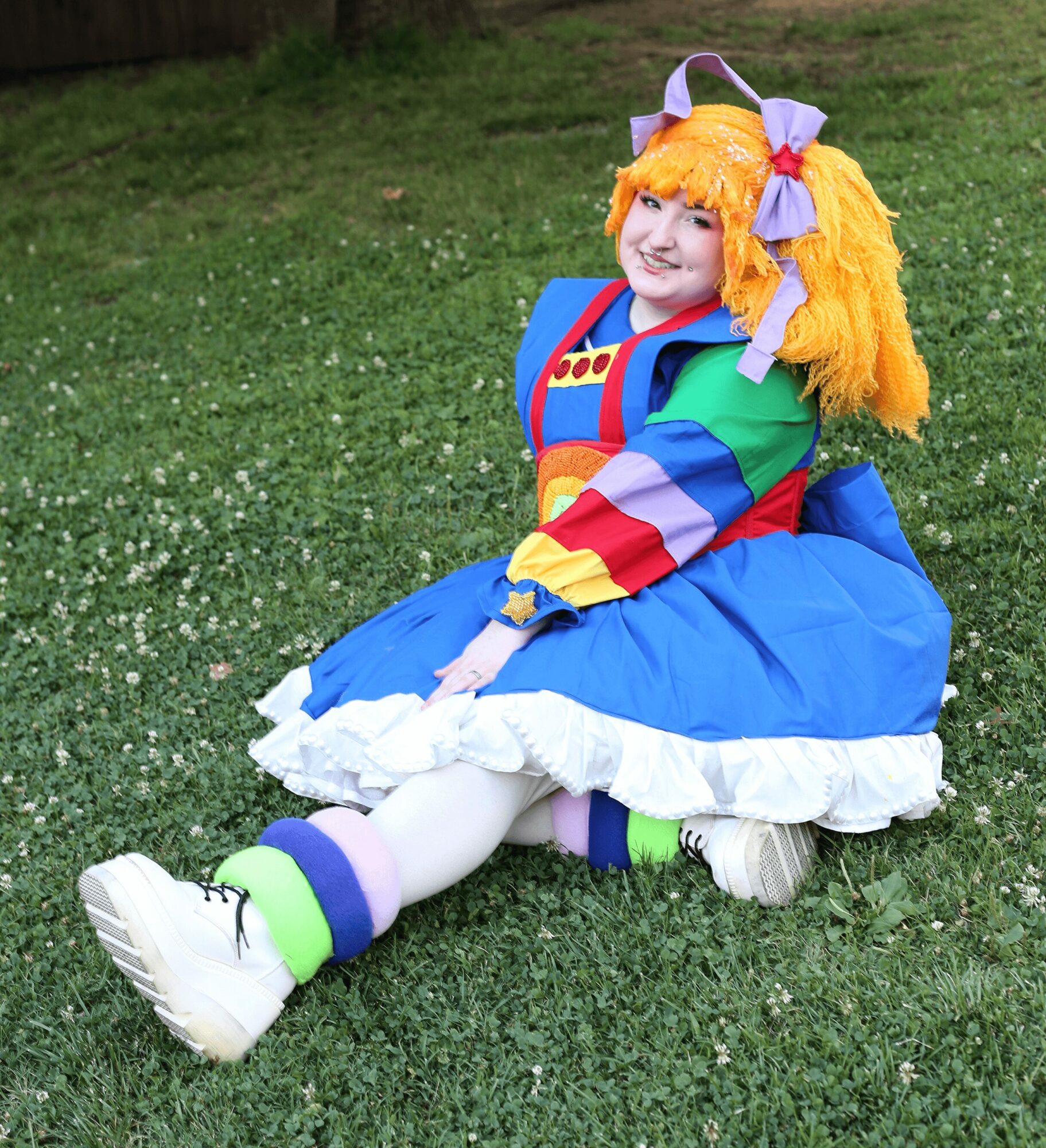 Person in colorful clown costume sitting on grass, smiling, with orange hair, large shoes, and a bow in hair.