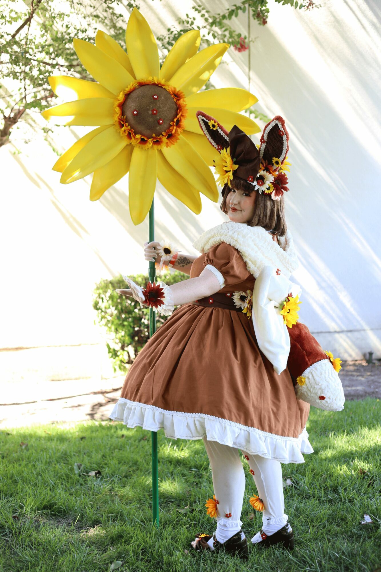 Child in costume with bunny ears, holding a sunflower umbrella, standing outdoors on grass with trees and sunlight.