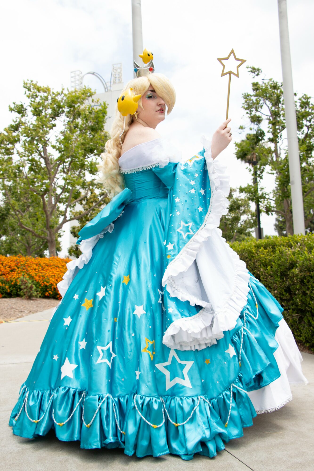 Person dressed as a fairy tale princess in a blue gown with star patterns, holding a star wand outdoors, trees in background.