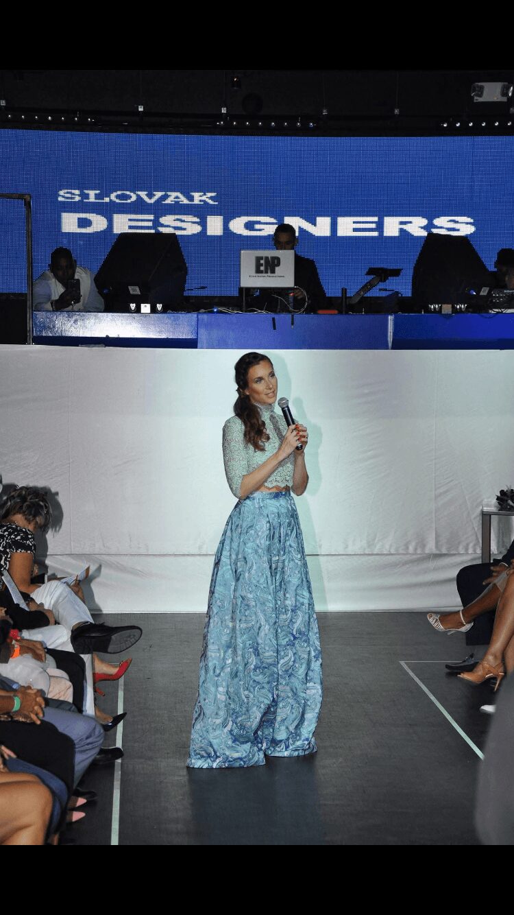 A woman in a blue patterned dress speaks into a microphone on stage at Slovakia Designers event, audience seated nearby.