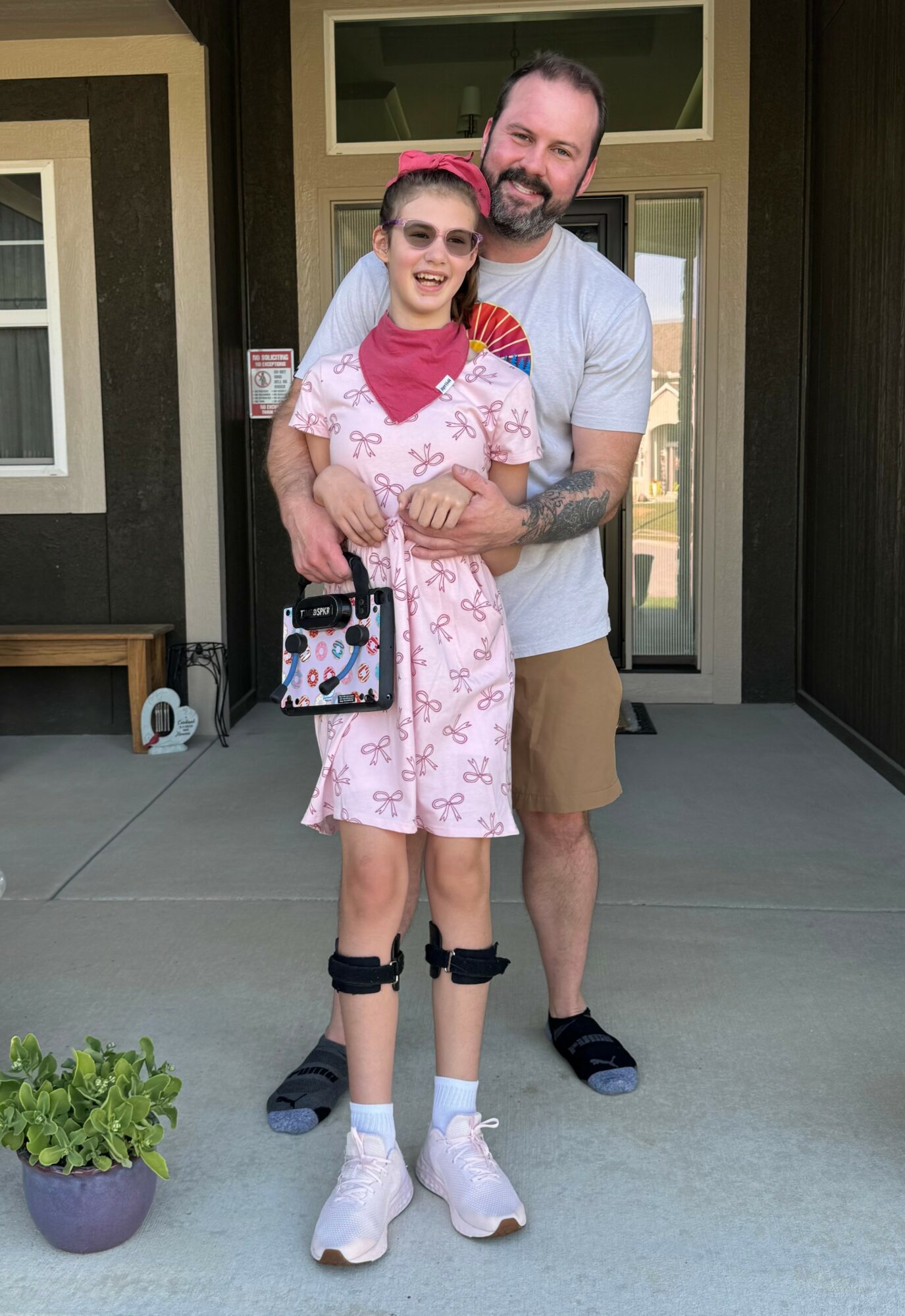 A man and girl stand outside a house, smiling, with the girl wearing sunglasses and a pink dress, man in a gray shirt and shorts.