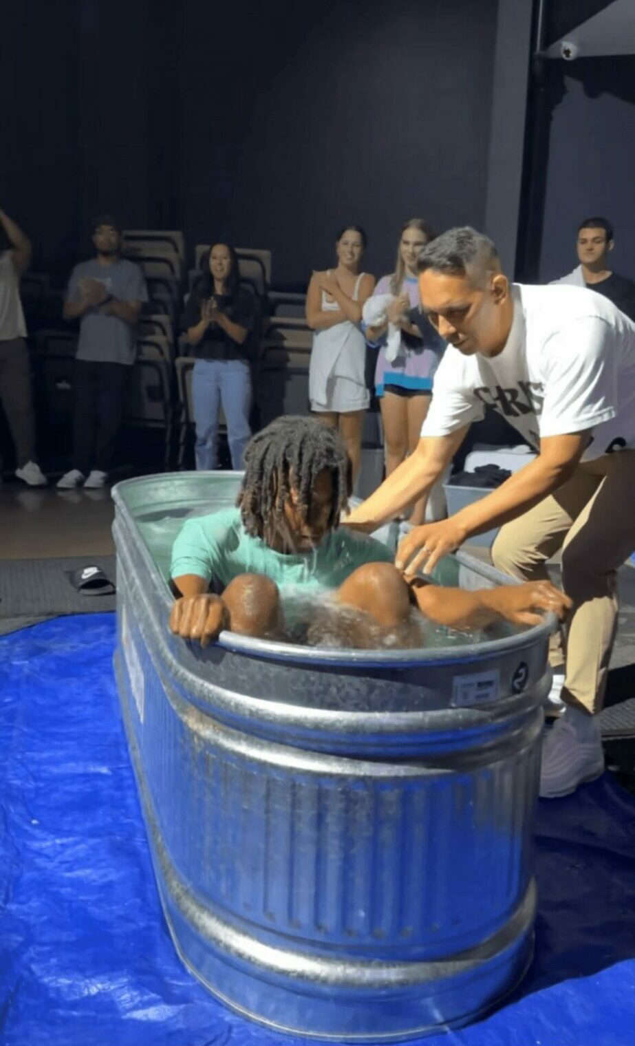 Person helping another in a metal tub, with onlookers in background, on a blue tarp.