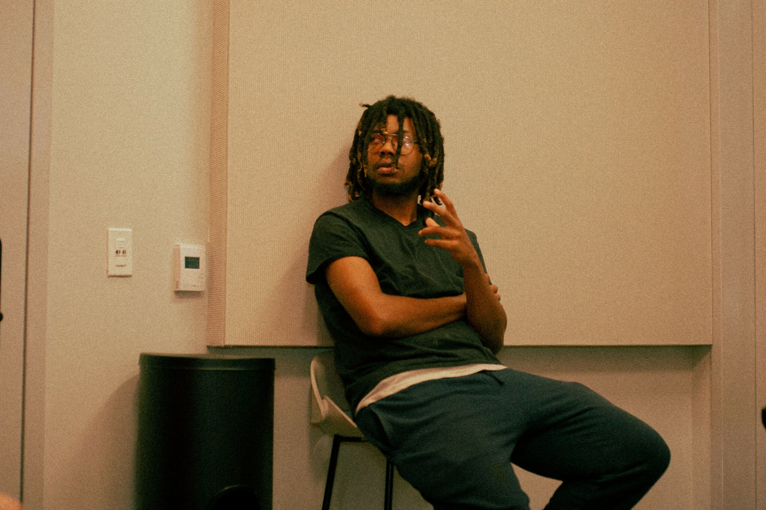 Person with dreadlocks sitting on a chair, taking a mirror selfie in a room with beige walls.