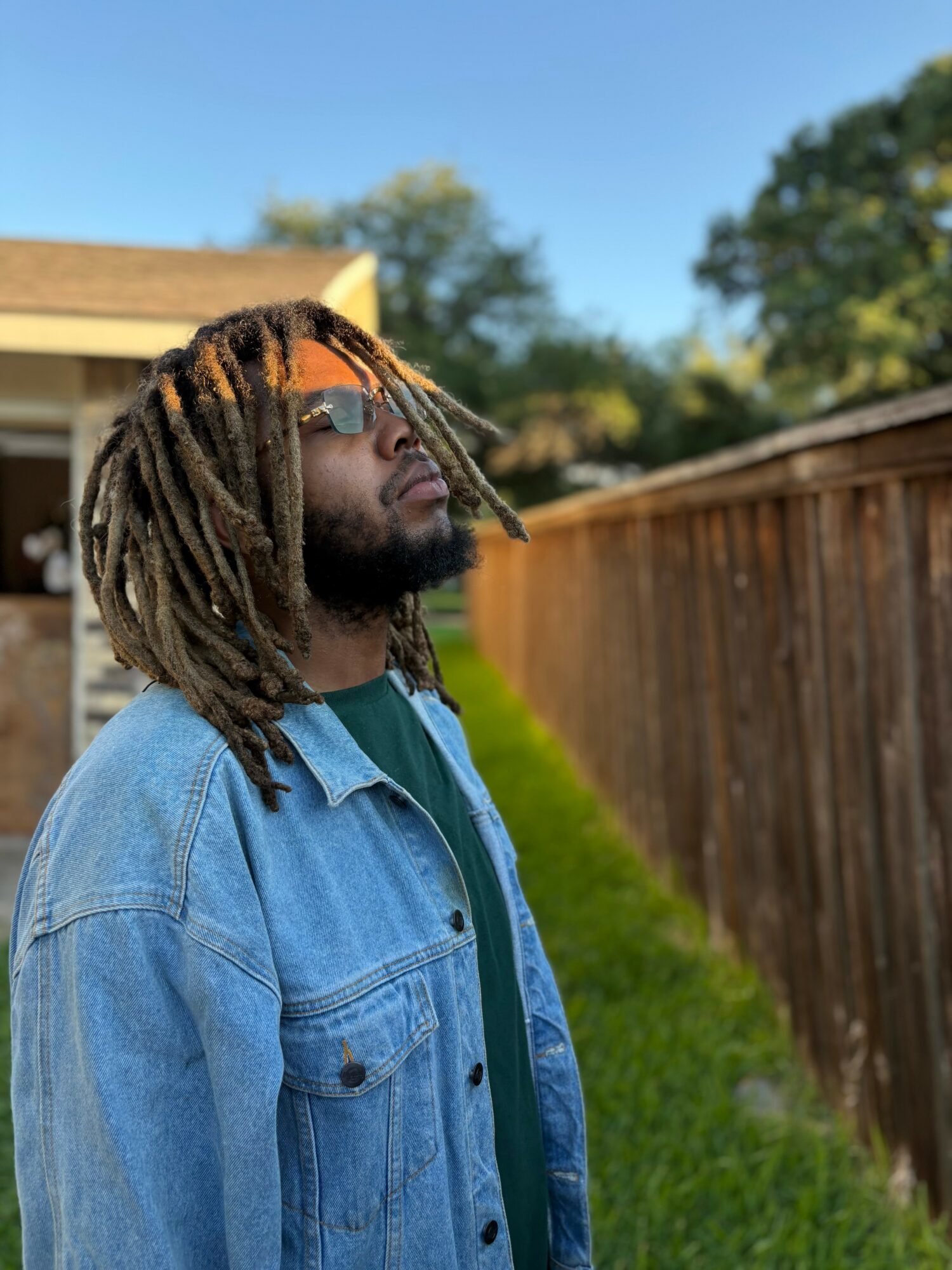 Young man with dreadlocks wearing sunglasses and a denim jacket outdoors, standing near a wooden fence, with trees and a house in background.