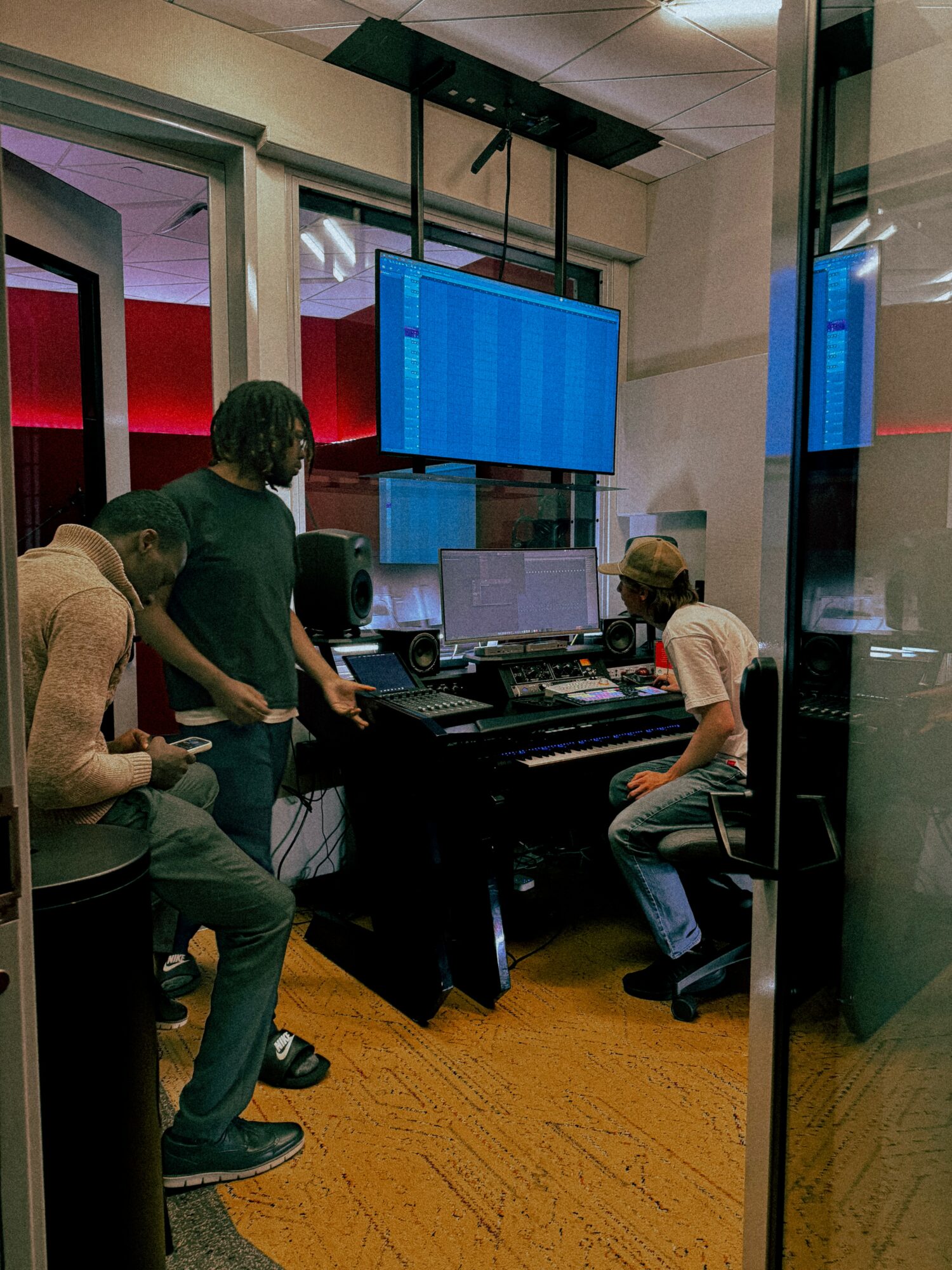 Four people in a recording studio with multiple monitors and a large screen, engaged in work or discussion.