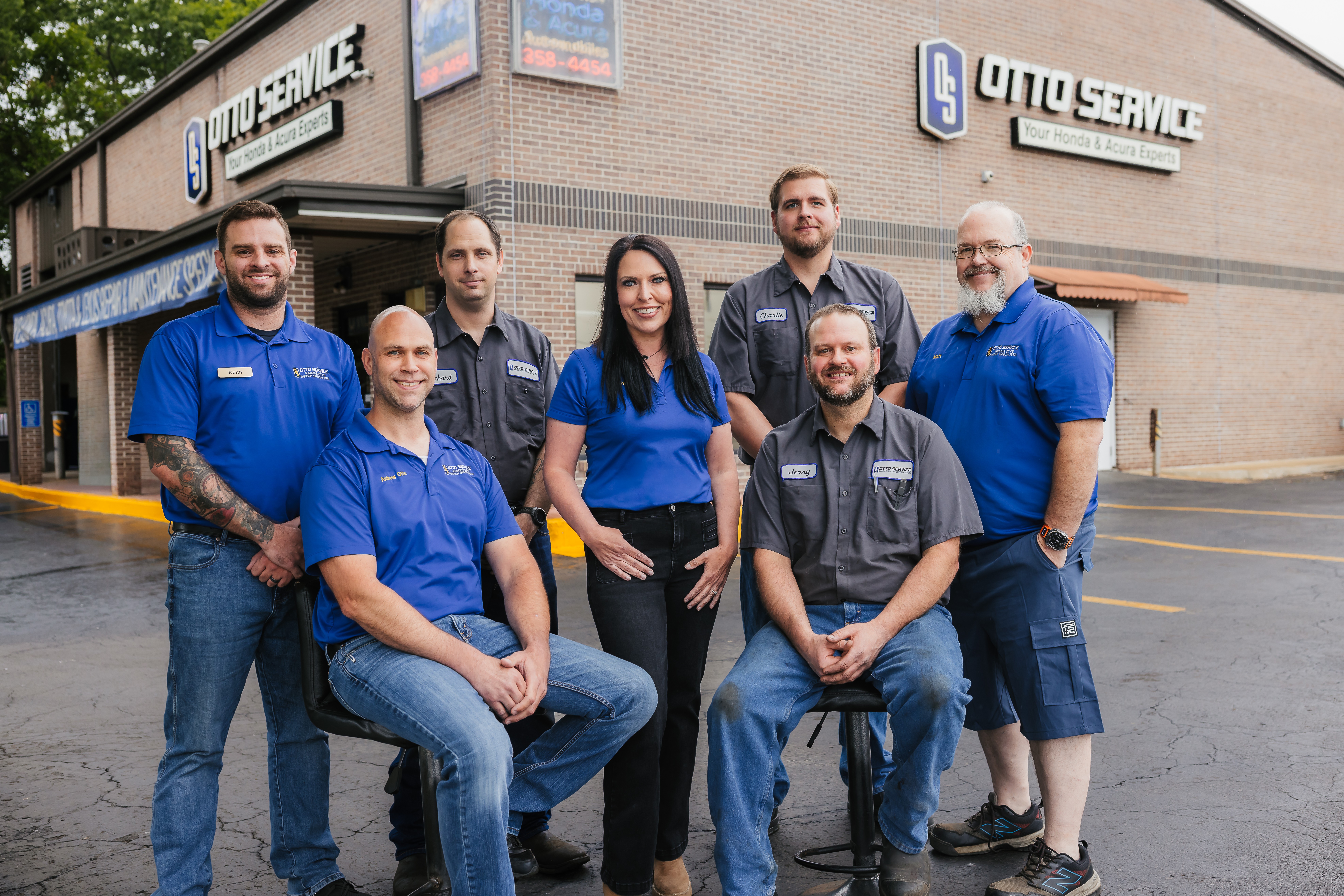 Group of seven people standing and sitting outside auto service building, smiling, wearing blue and black uniforms.