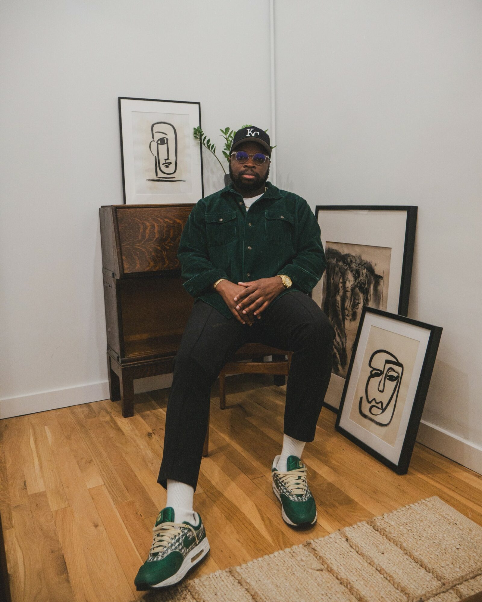 Person sitting on a chair in an art gallery, surrounded by framed artwork on the floor and wall, hardwood floor, white wall.