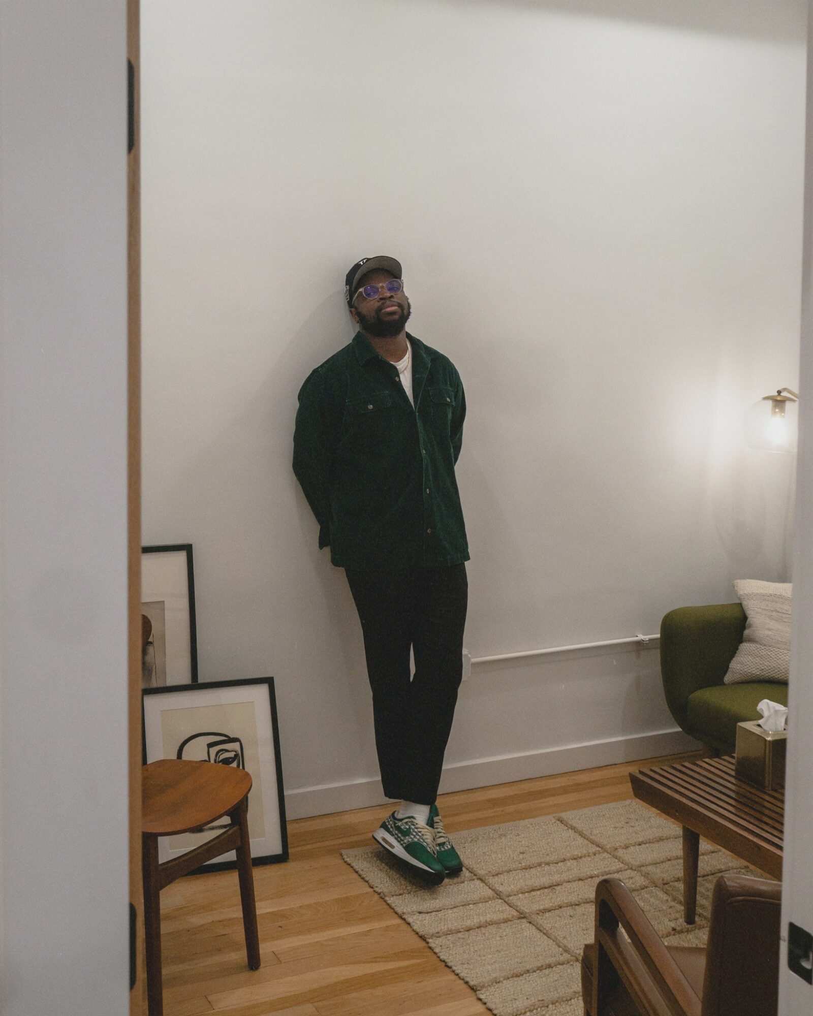 Person standing against a white wall, wearing dark clothing and sneakers, with framed pictures on the floor nearby.