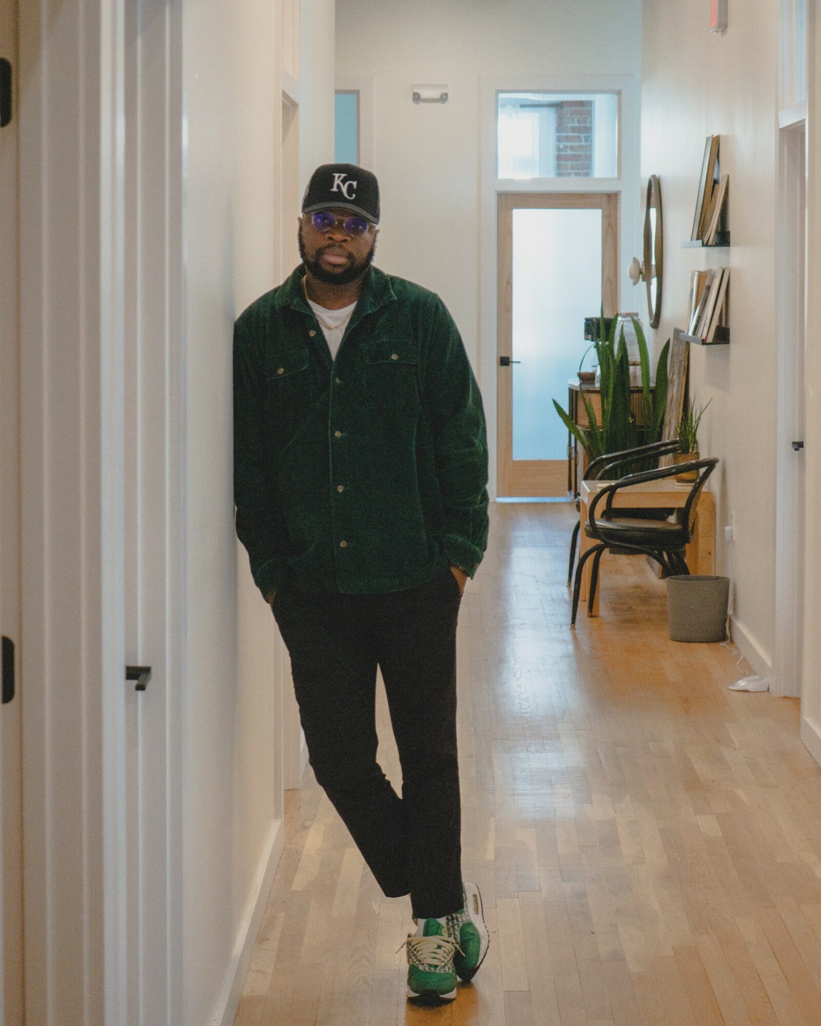 Man standing in a hallway with wooden flooring, white walls, and furniture, wearing a black jacket, cap, and sneakers.