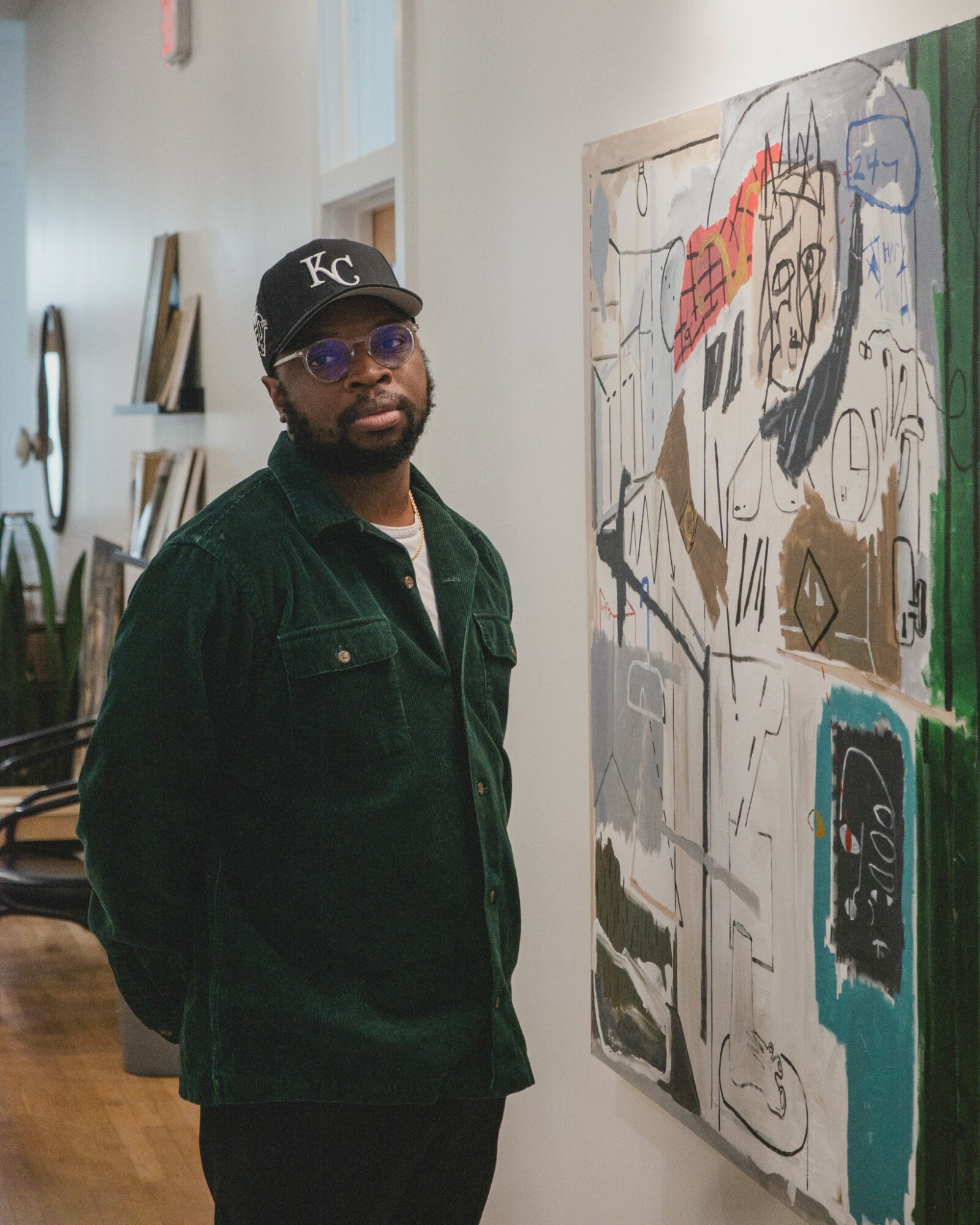 Man wearing a black cap, sunglasses, and dark jacket standing next to abstract artwork in gallery.