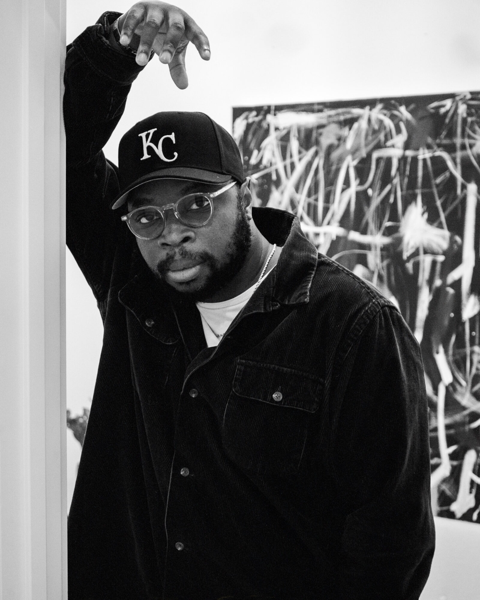 Man wearing glasses and a Kansas City baseball cap, raising his hand, standing in front of abstract artwork.