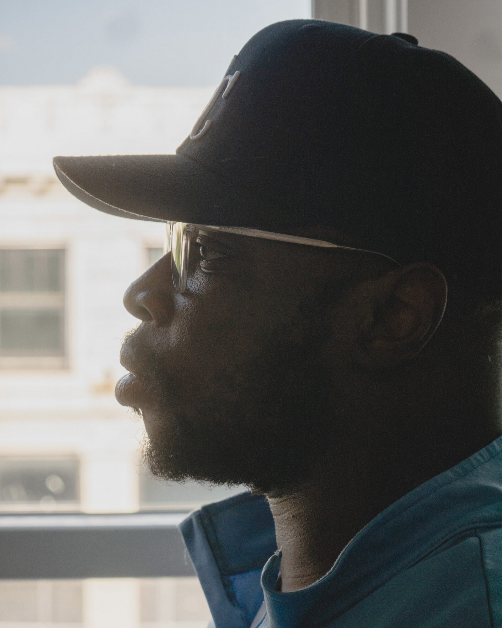 Profile of a person wearing a cap and glasses, looking out a window with buildings outside.