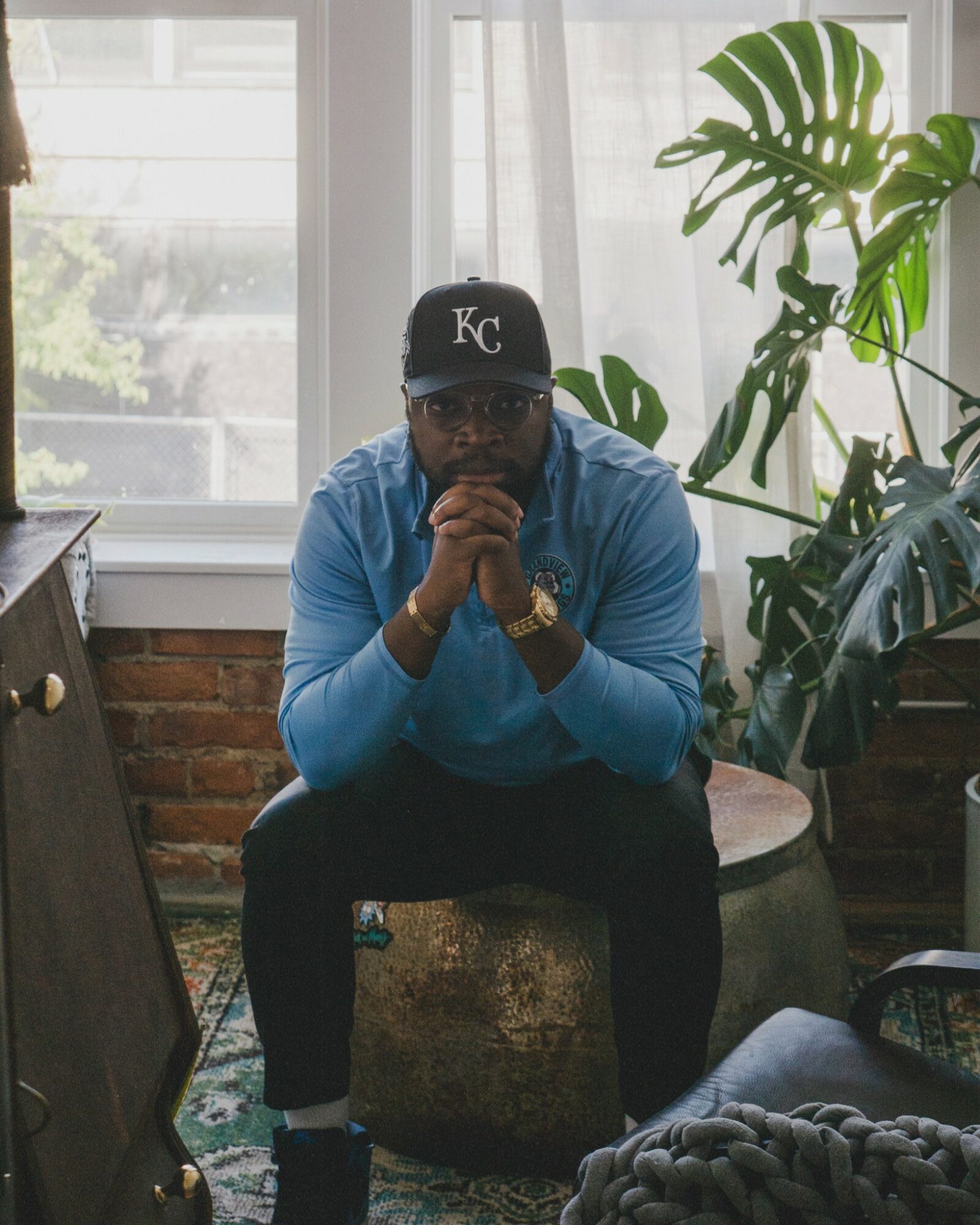 Person sitting on a round ottoman with hands clasped, wearing a black cap, blue hoodie, and glasses, in a room with large windows and plants.