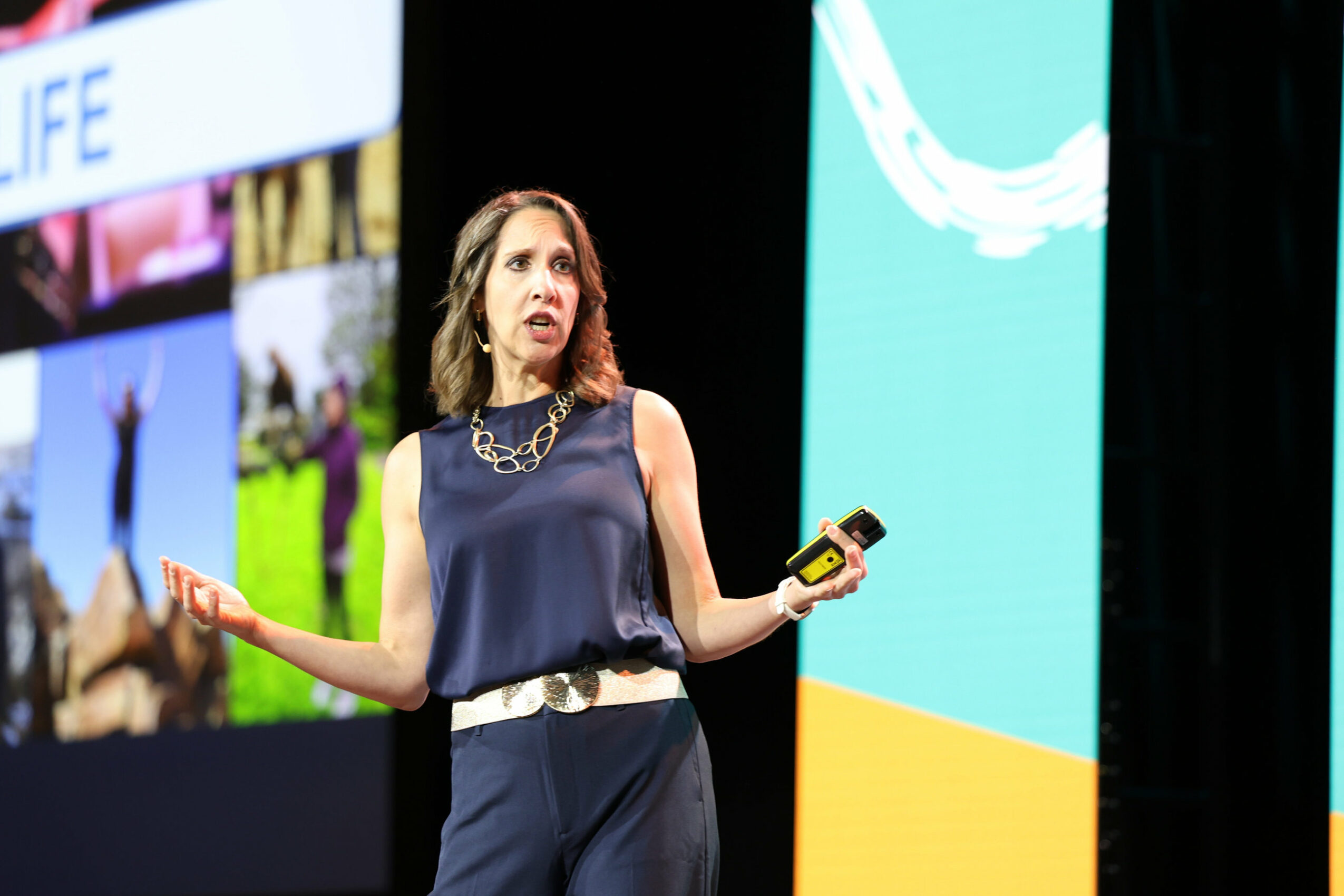 Woman speaking on stage holding a microphone and a remote, with a large screen behind her displaying colorful graphics.