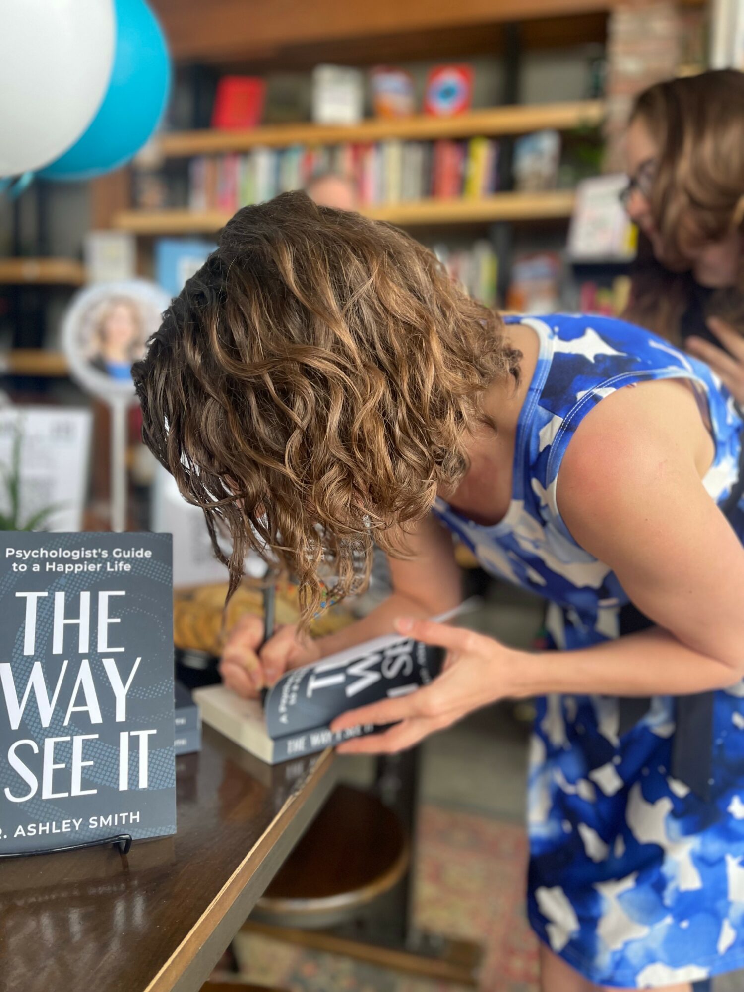 Person with curly hair reads a book at a table in a bookstore or library, with shelves of books in the background.