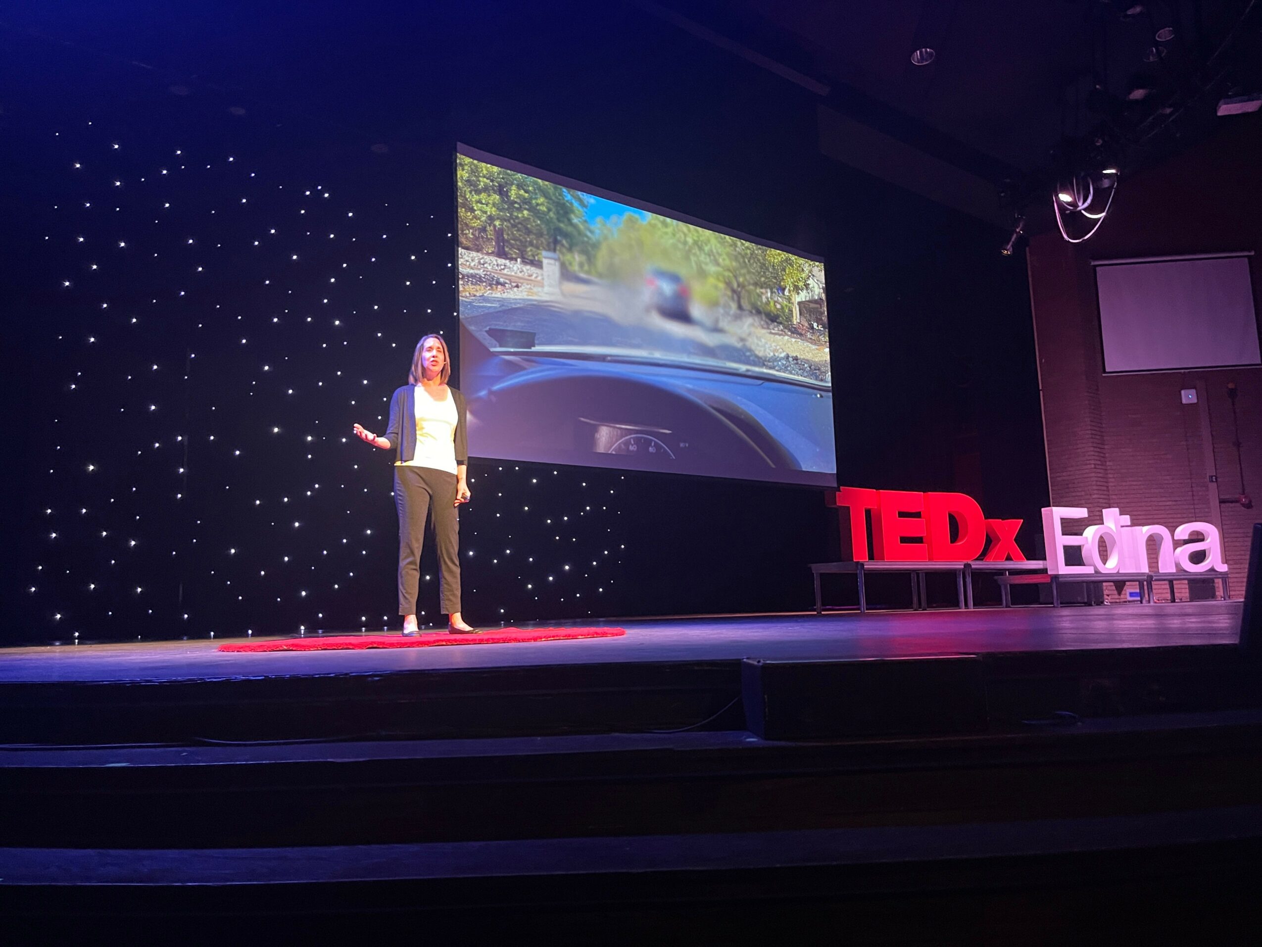 Woman standing on stage with a large screen behind her displaying a road view, TEDx Edna sign on stage, dark background.