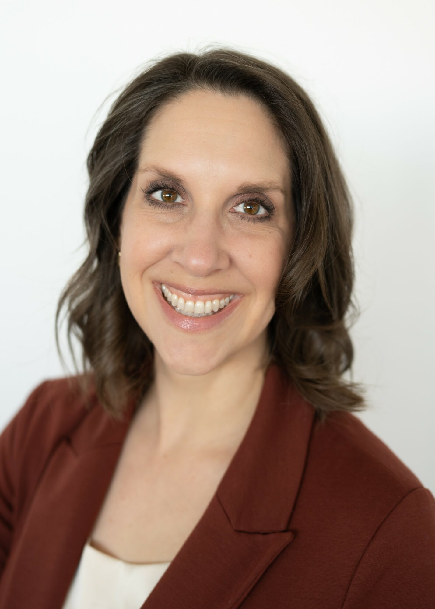 Smiling woman with shoulder-length wavy hair, wearing a brown blazer and white top, against a plain white background.