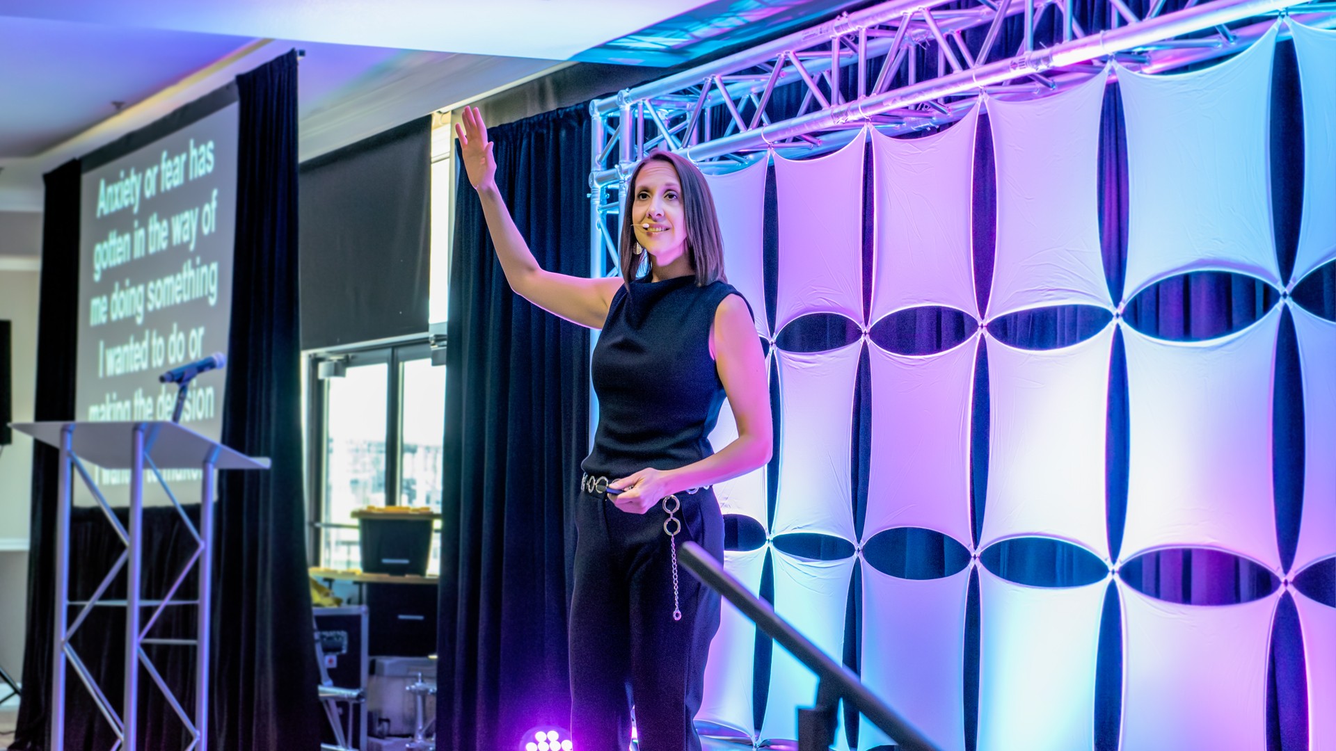 Woman on stage gesturing with her right hand, background with geometric patterns and stage lighting, conference setting.