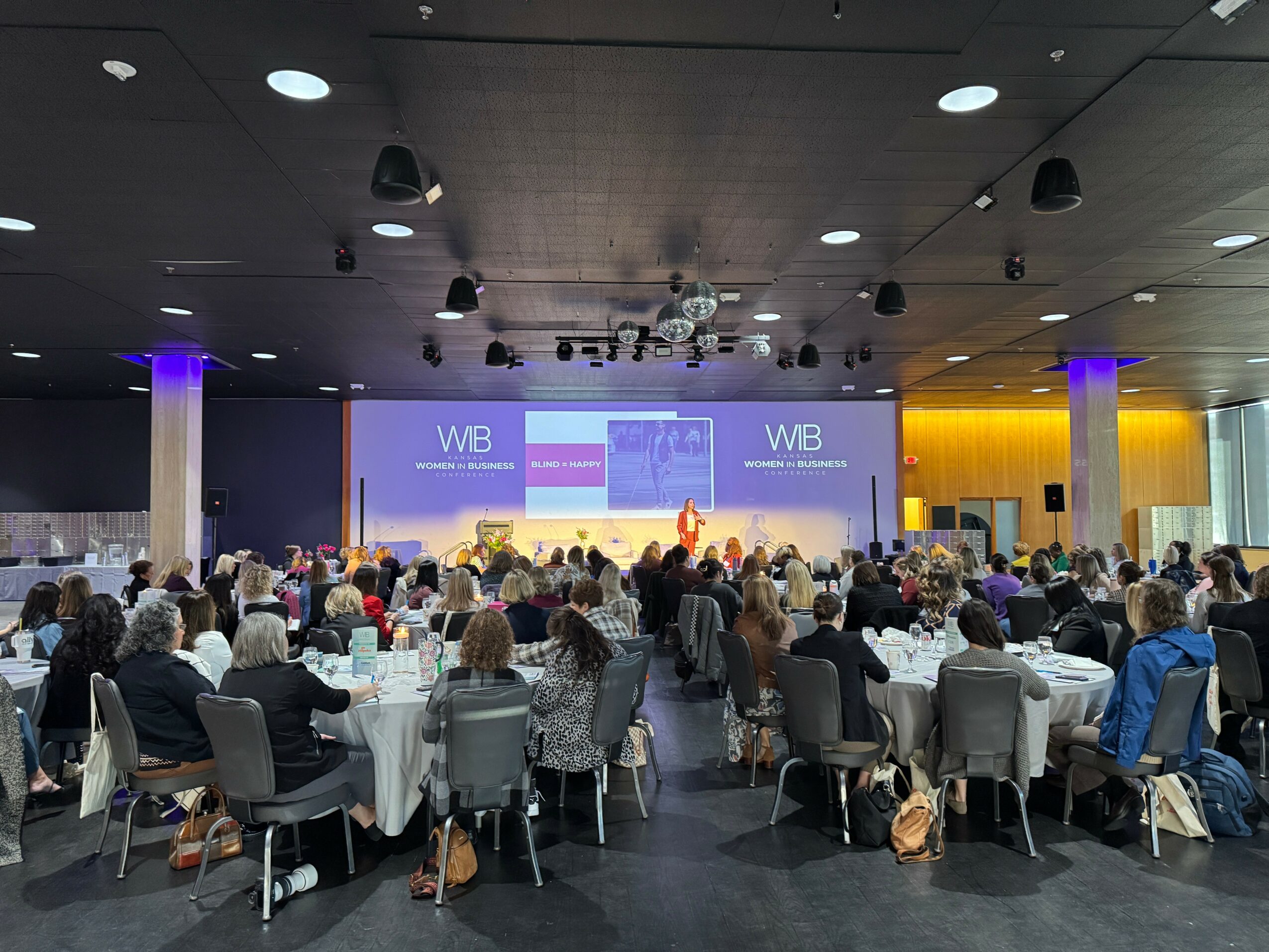 Conference room filled with seated attendees facing a stage with large screens displaying presentations.