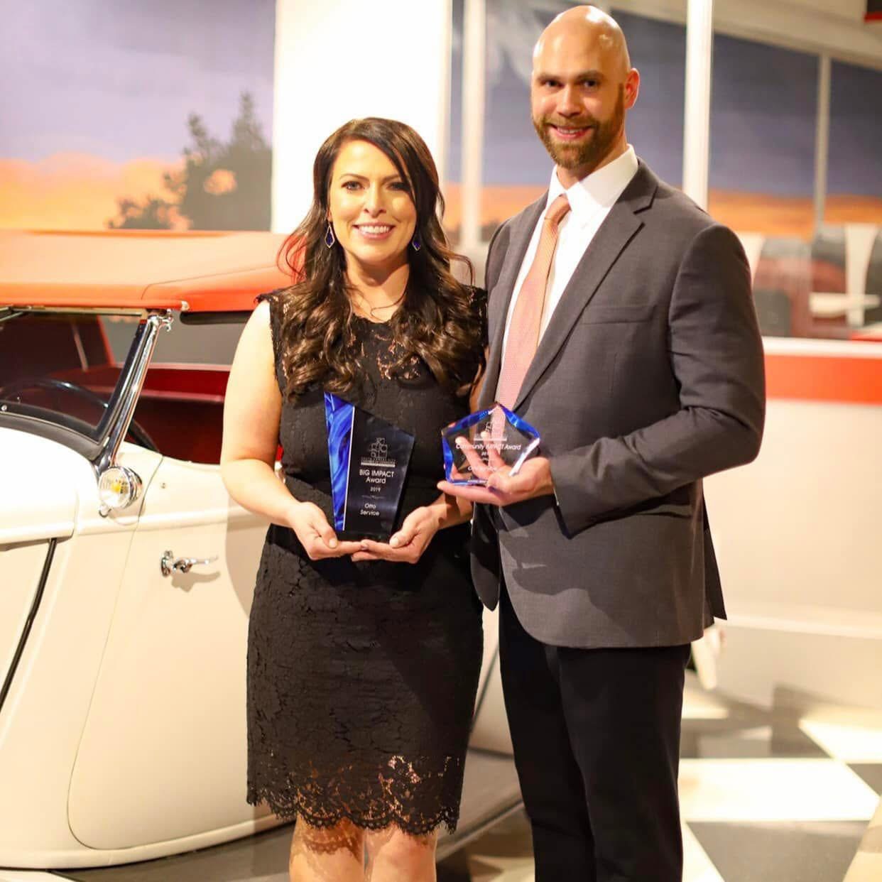 A woman and a man in formal attire holding awards, standing indoors near a vintage car, with sunset scenery outside.
