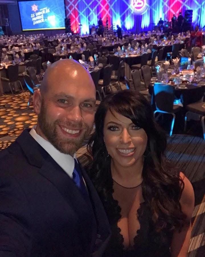 Two people smiling at a formal event with tables and a stage in the background.