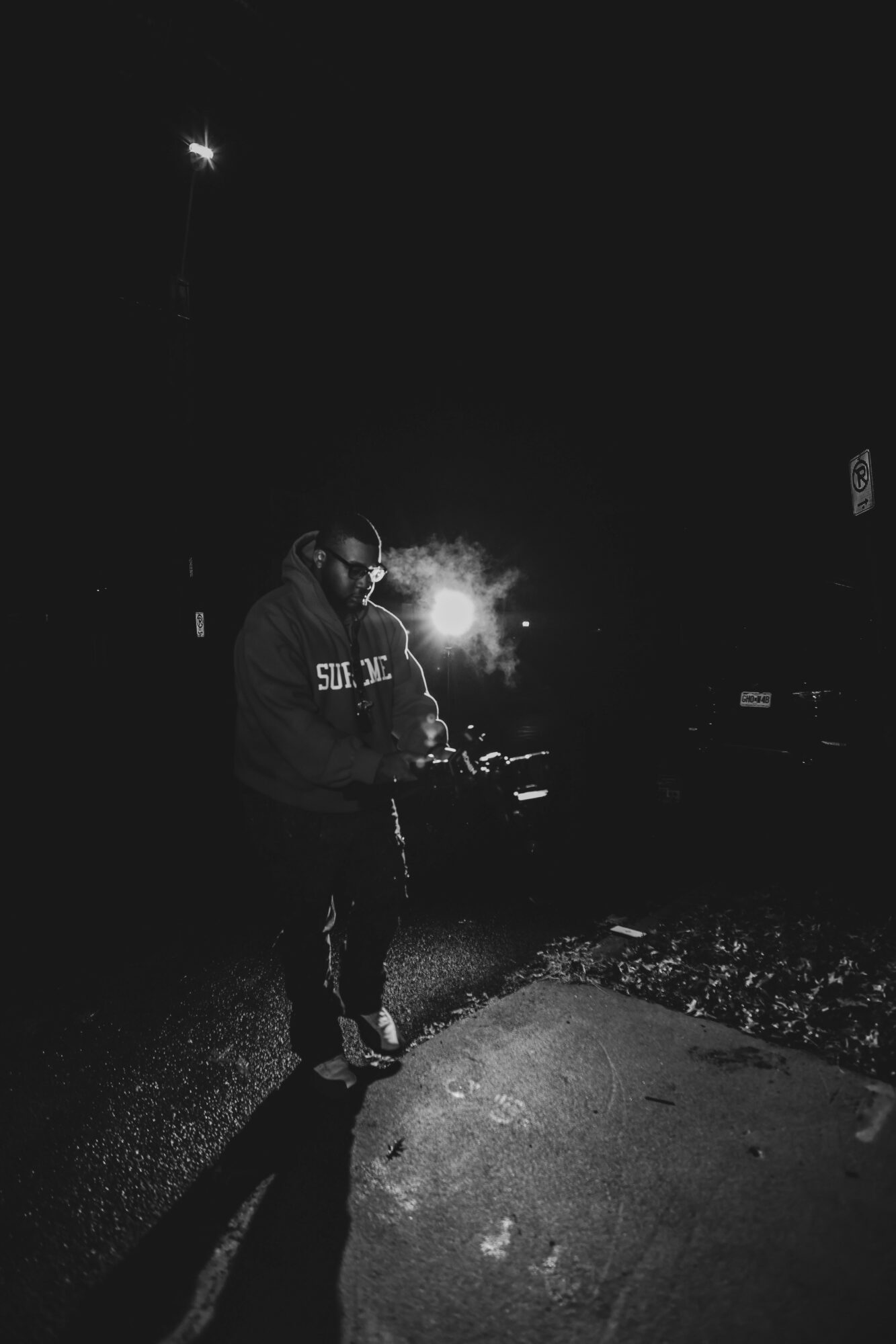 Person standing outdoors at night, illuminated by a bright light behind them, with a dark background and street elements.