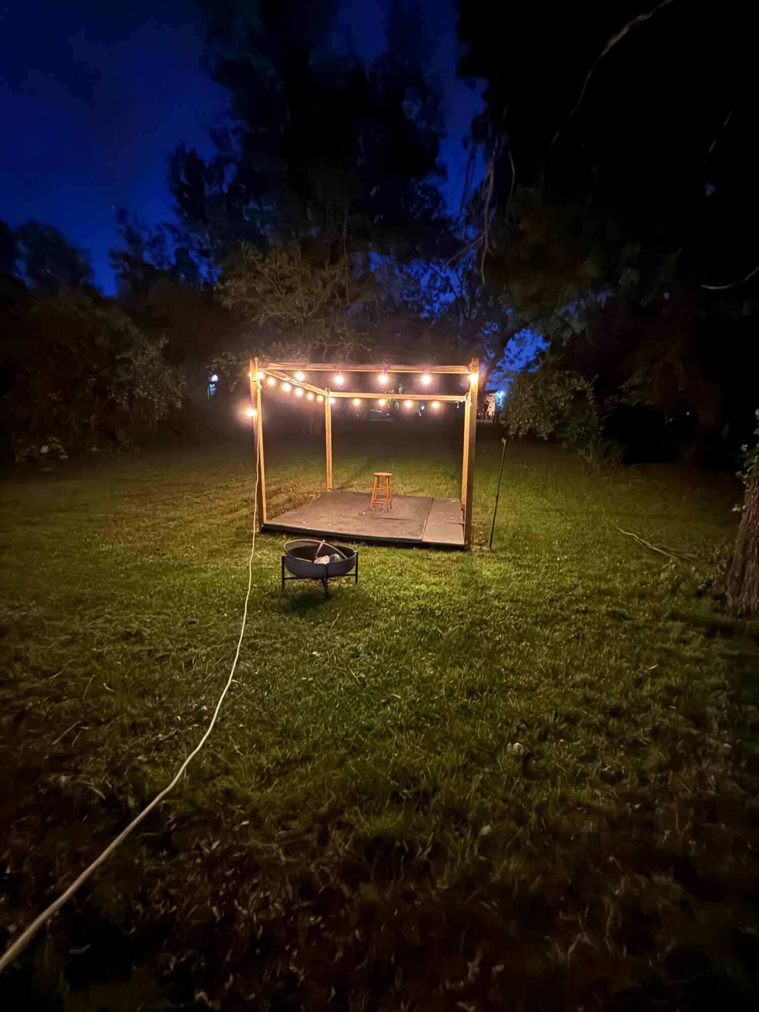 A wooden frame with string lights stands on grass at night, with trees and a dark sky in the background.