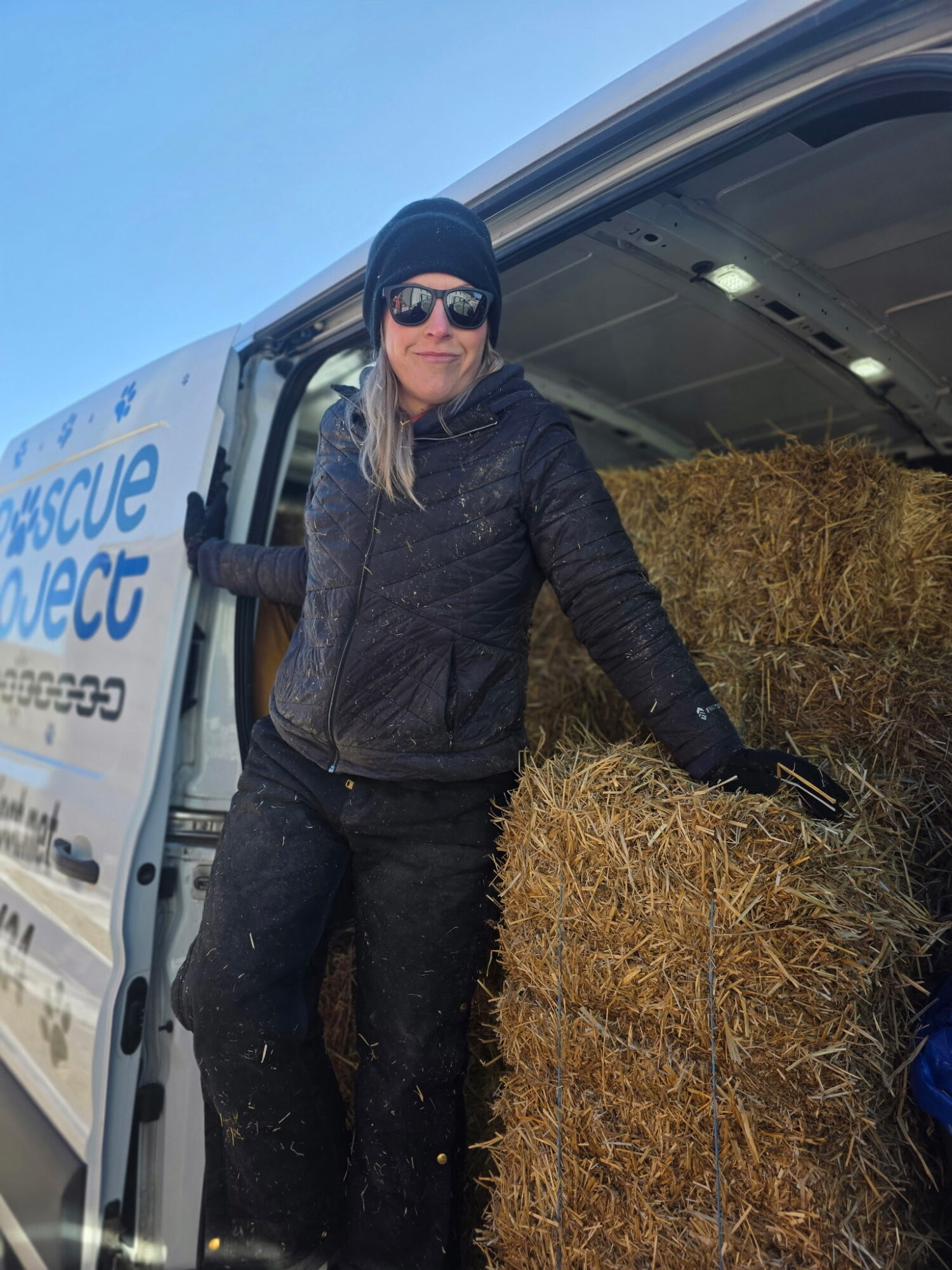 Person wearing sunglasses, black beanie, black jacket, standing next to hay bales in a vehicle, outdoors with blue sky.