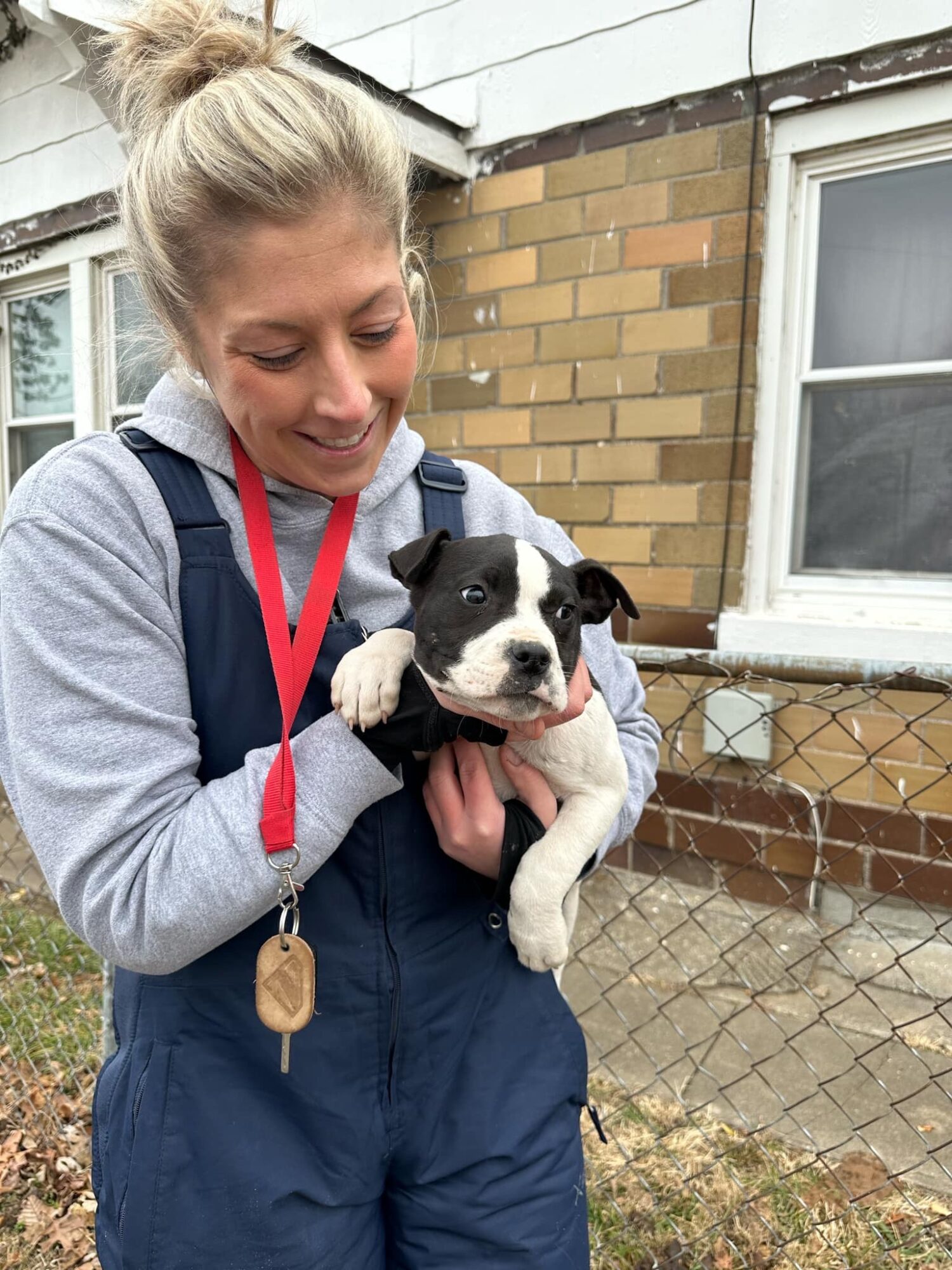 Woman holding a small black and white puppy outside near a brick building and window.