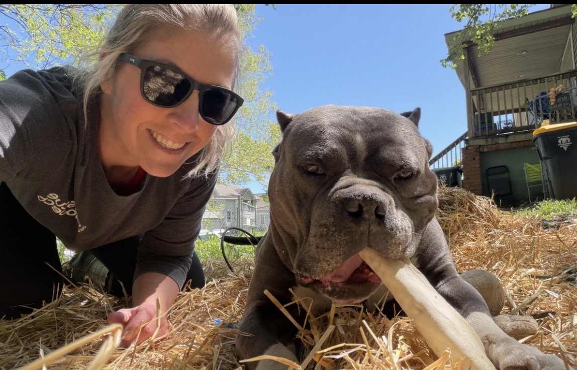 Woman with sunglasses smiling outdoors next to a large dog chewing a stick on dry grass.