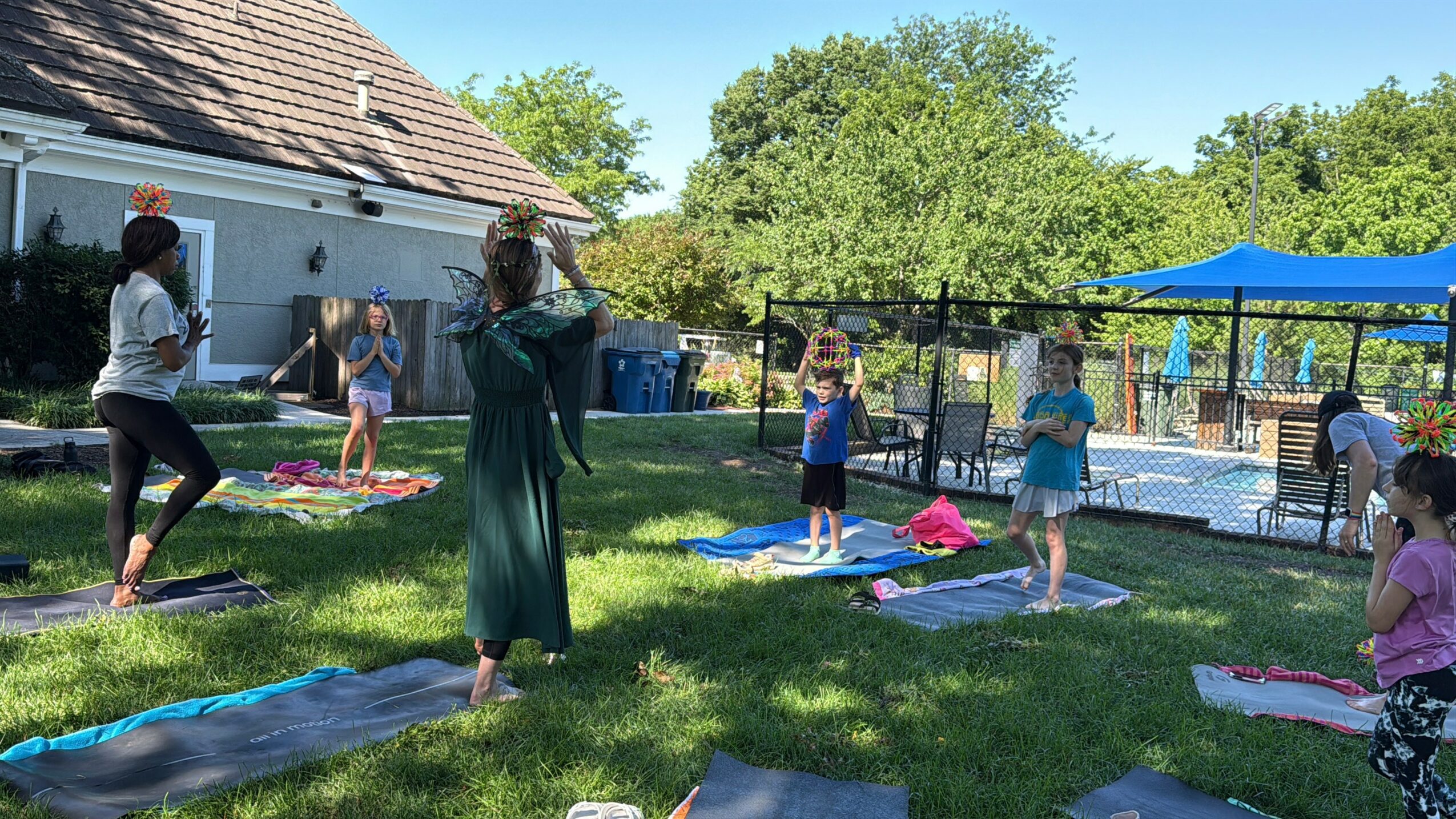 People participating in an outdoor yoga class on mats in a grassy backyard with trees, a house, and a pool in the background.