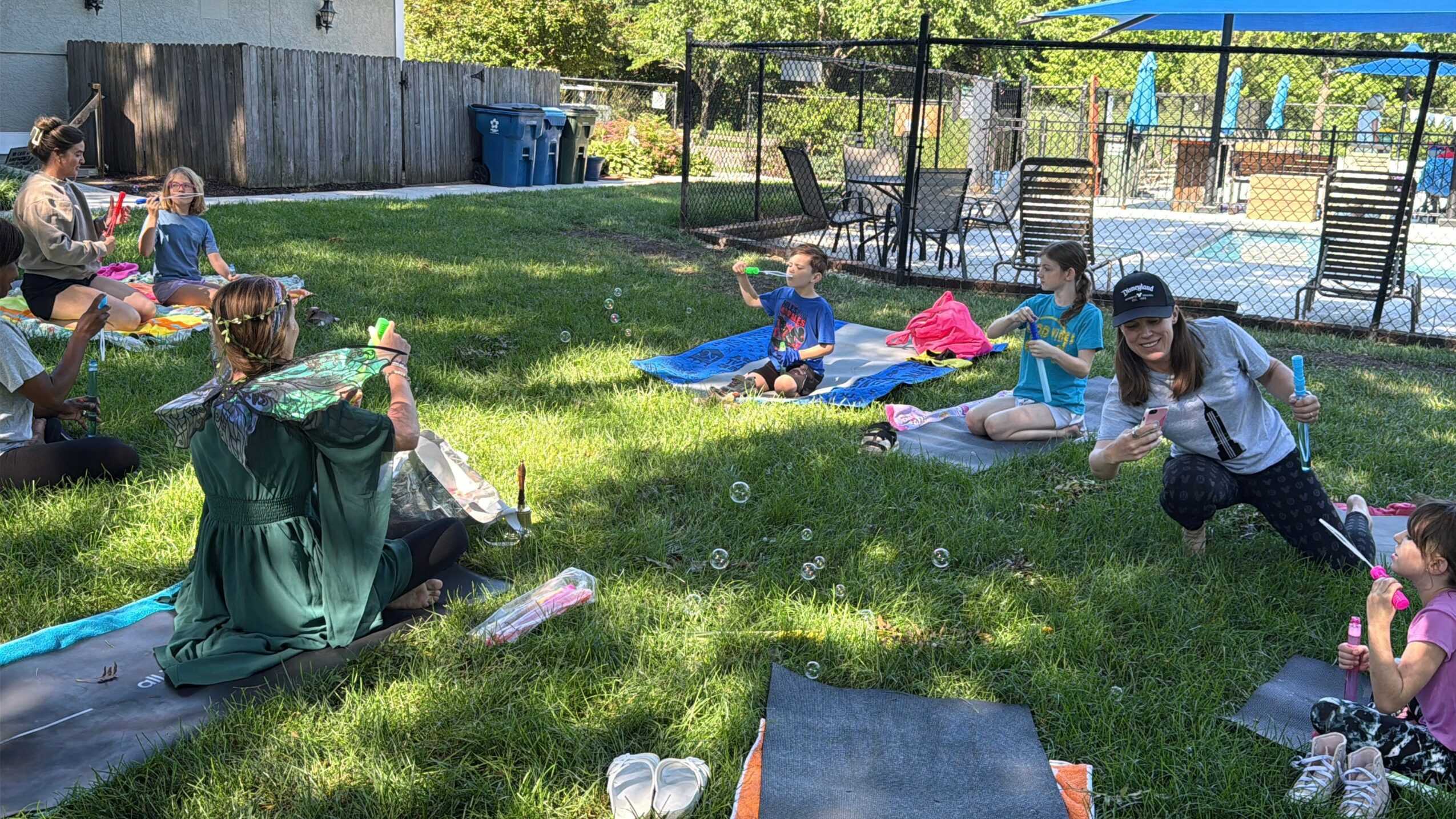Group of children and adults sitting on mats in a grassy backyard, engaging in outdoor activities.