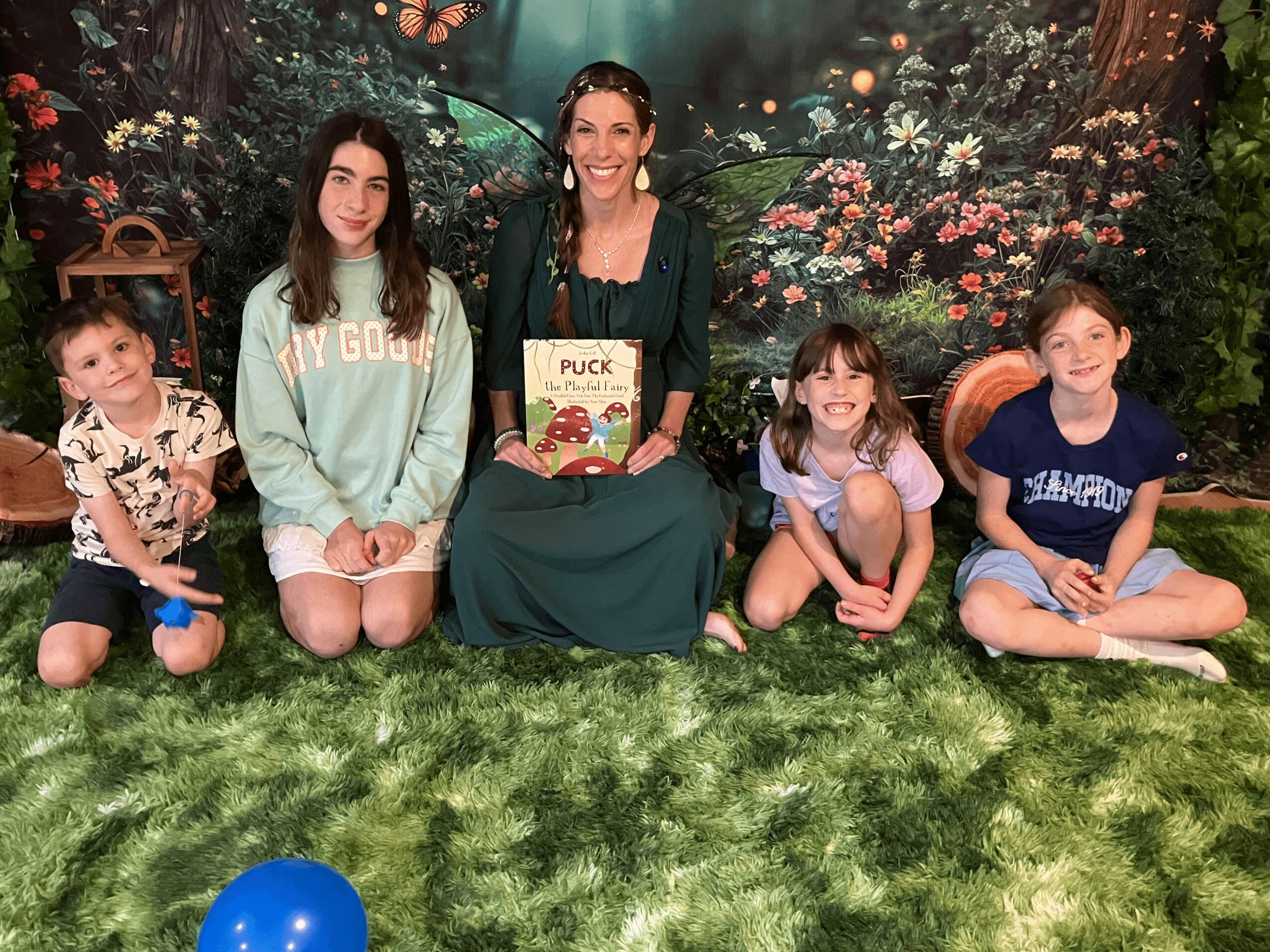 Group of five children and a woman sitting on grass in front of a forest backdrop, smiling, with a blue ball nearby.