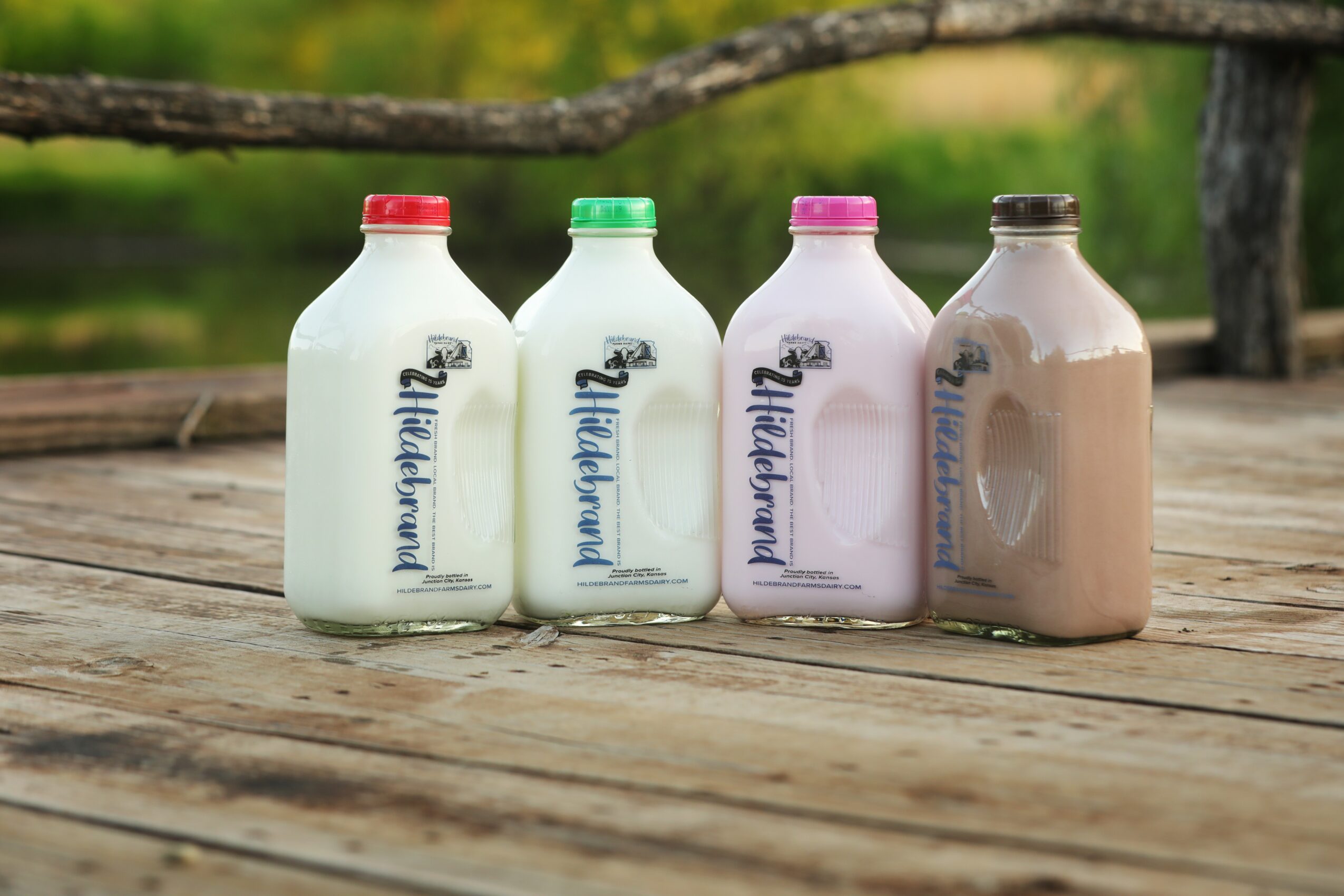 Four bottles of flavored milk on a wooden surface outdoors, with a blurred green background, each with different colored caps.