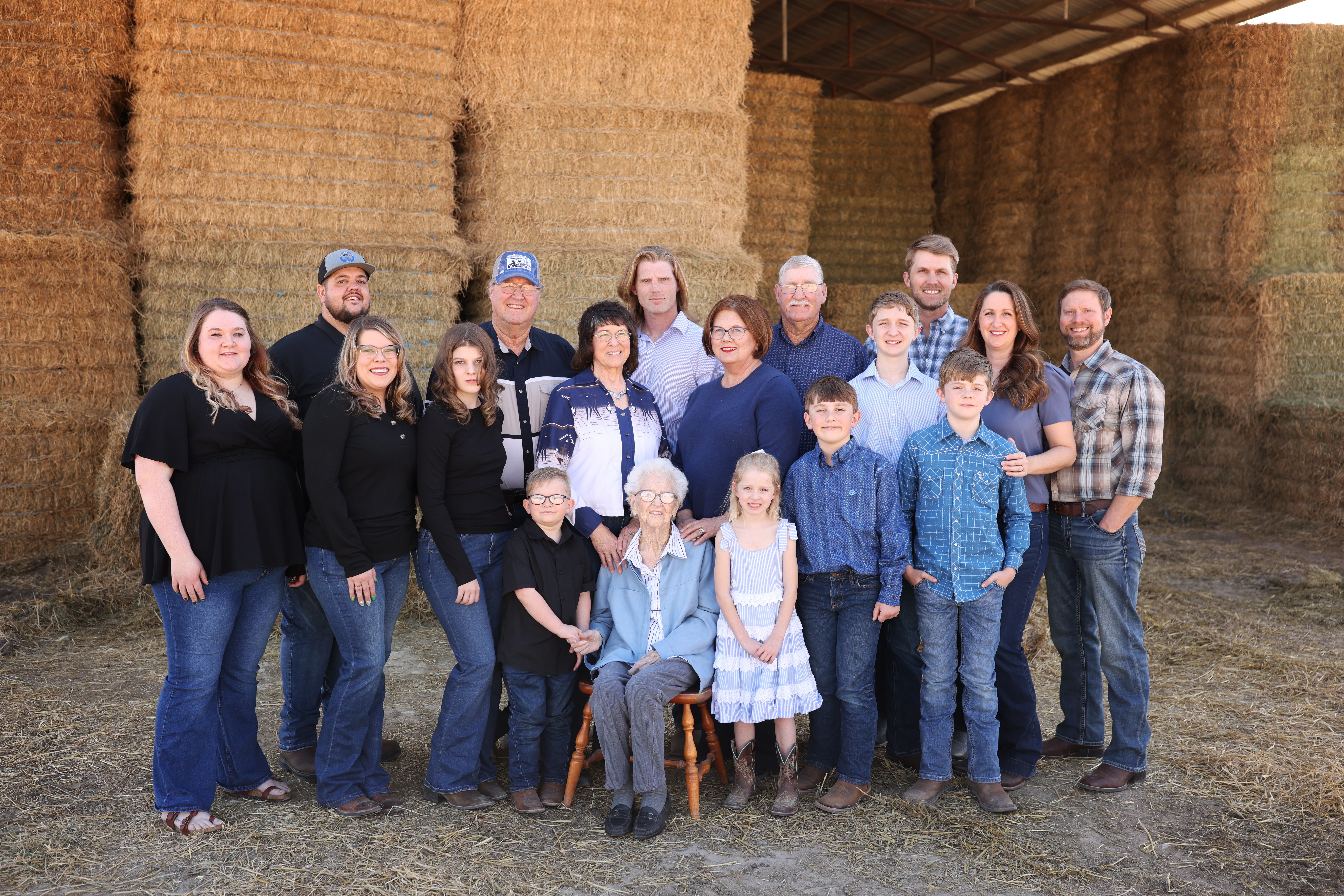 Group of 16 people standing inside a barn with hay bales, some smiling, dressed casually.