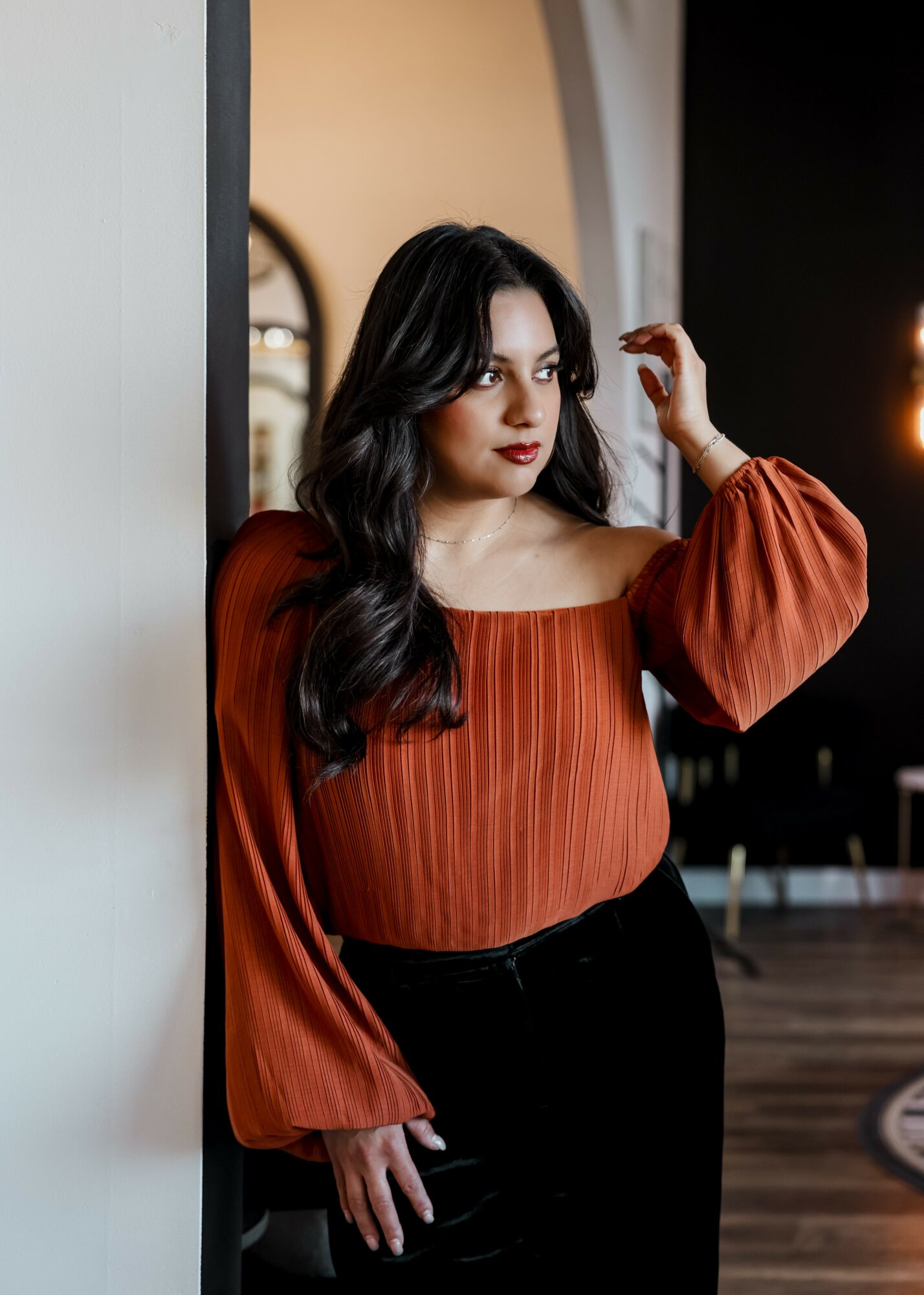 Woman with long dark hair wearing an off-shoulder orange top and black pants, standing indoors near a wall.