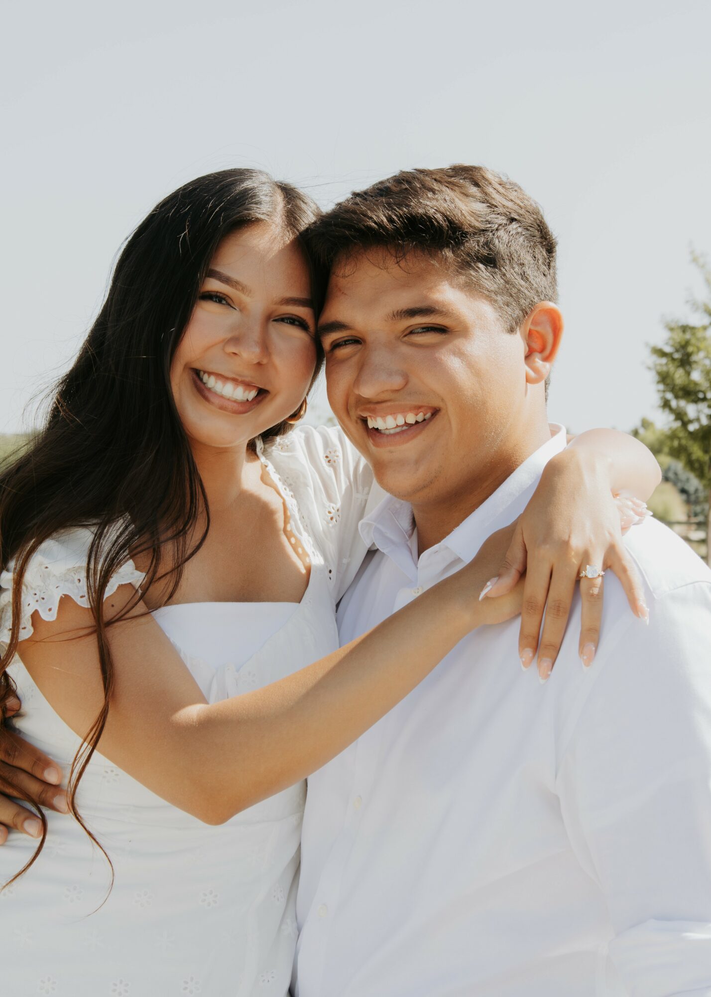 A smiling couple embraces outdoors, with the woman wearing a white dress and the man in a white shirt.