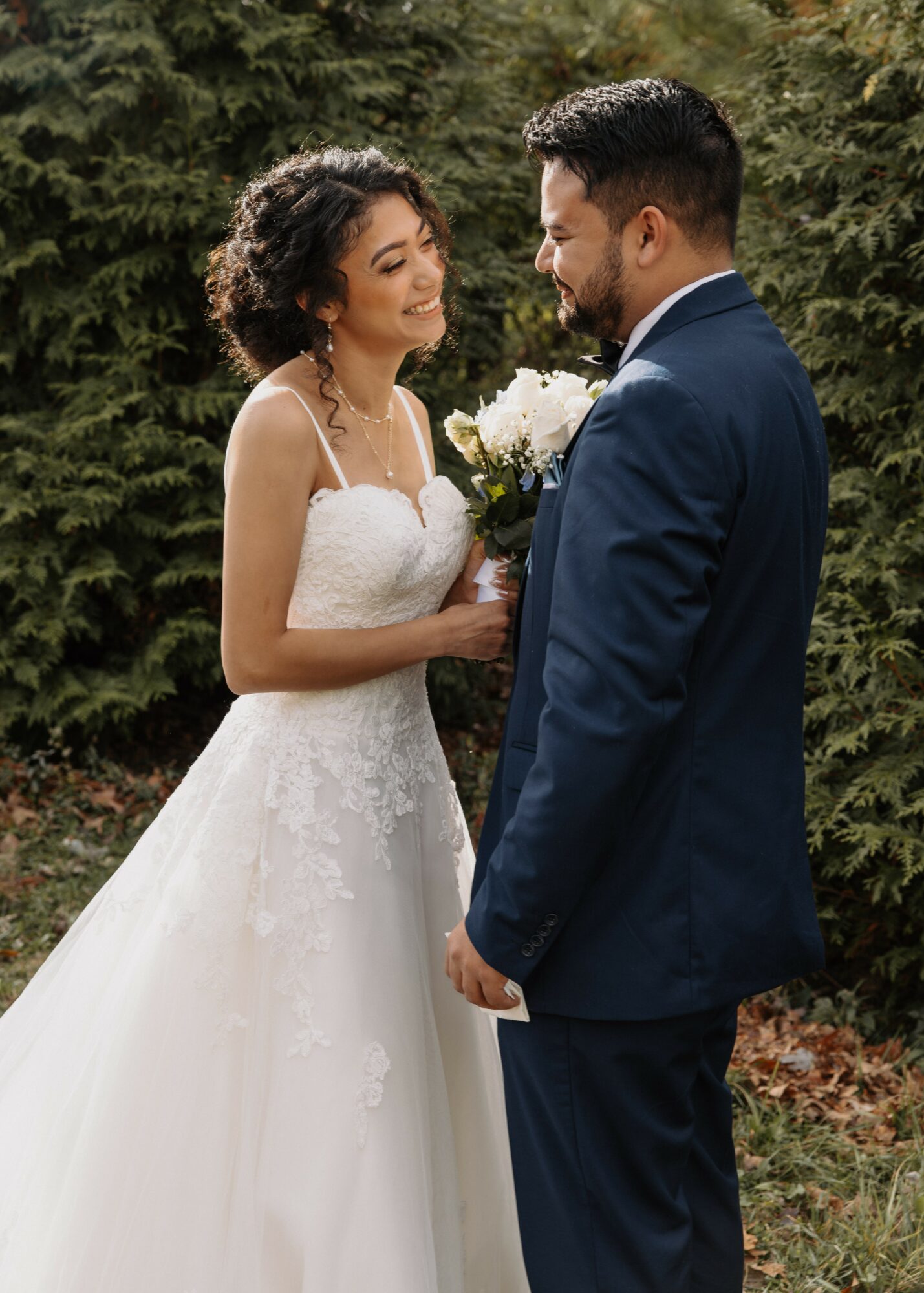 Bride and groom face each other outdoors, bride holding a bouquet, surrounded by greenery.