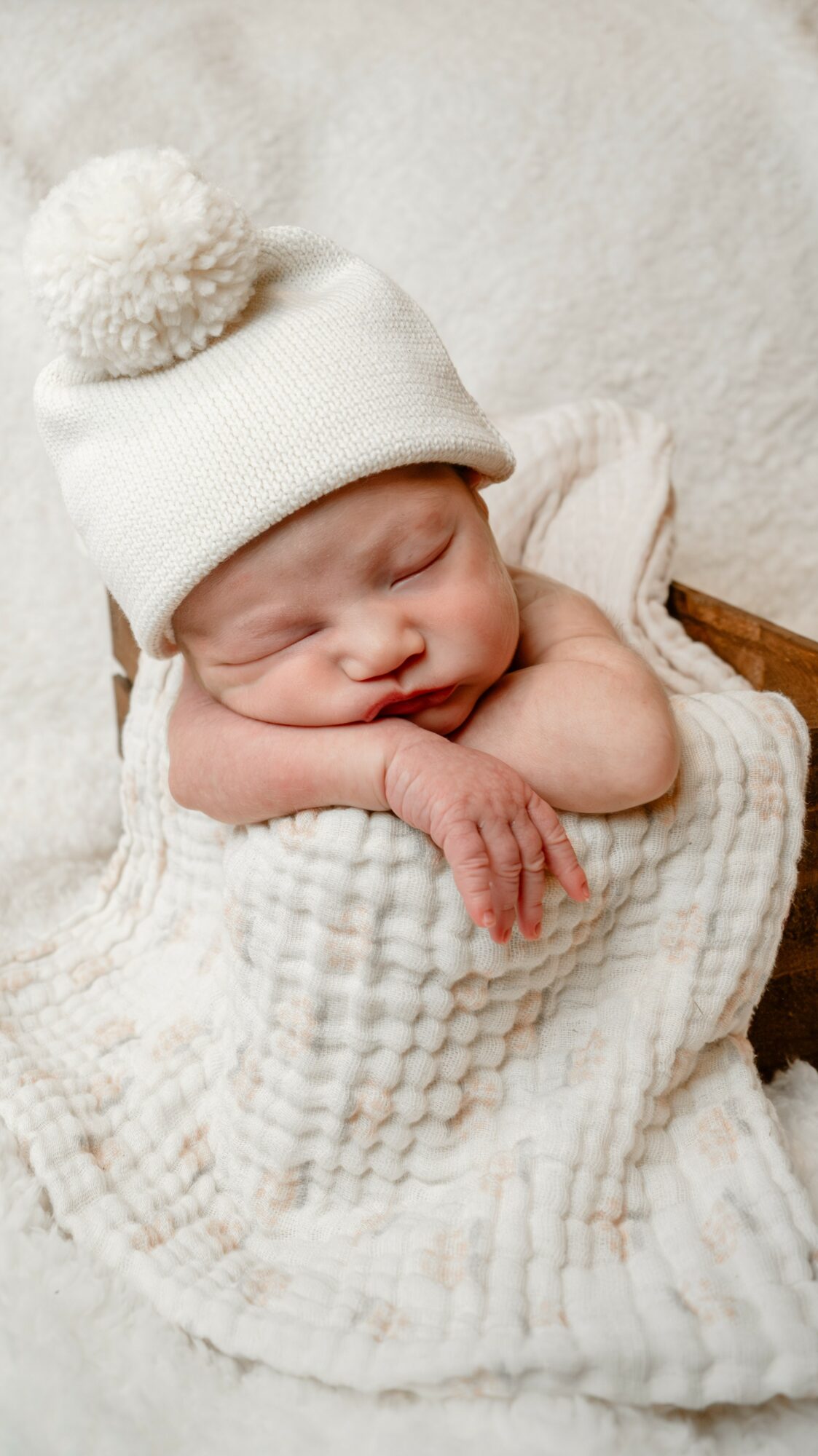Baby sleeping with head resting on hands, wearing a white knit hat with pom-pom, wrapped in a soft blanket