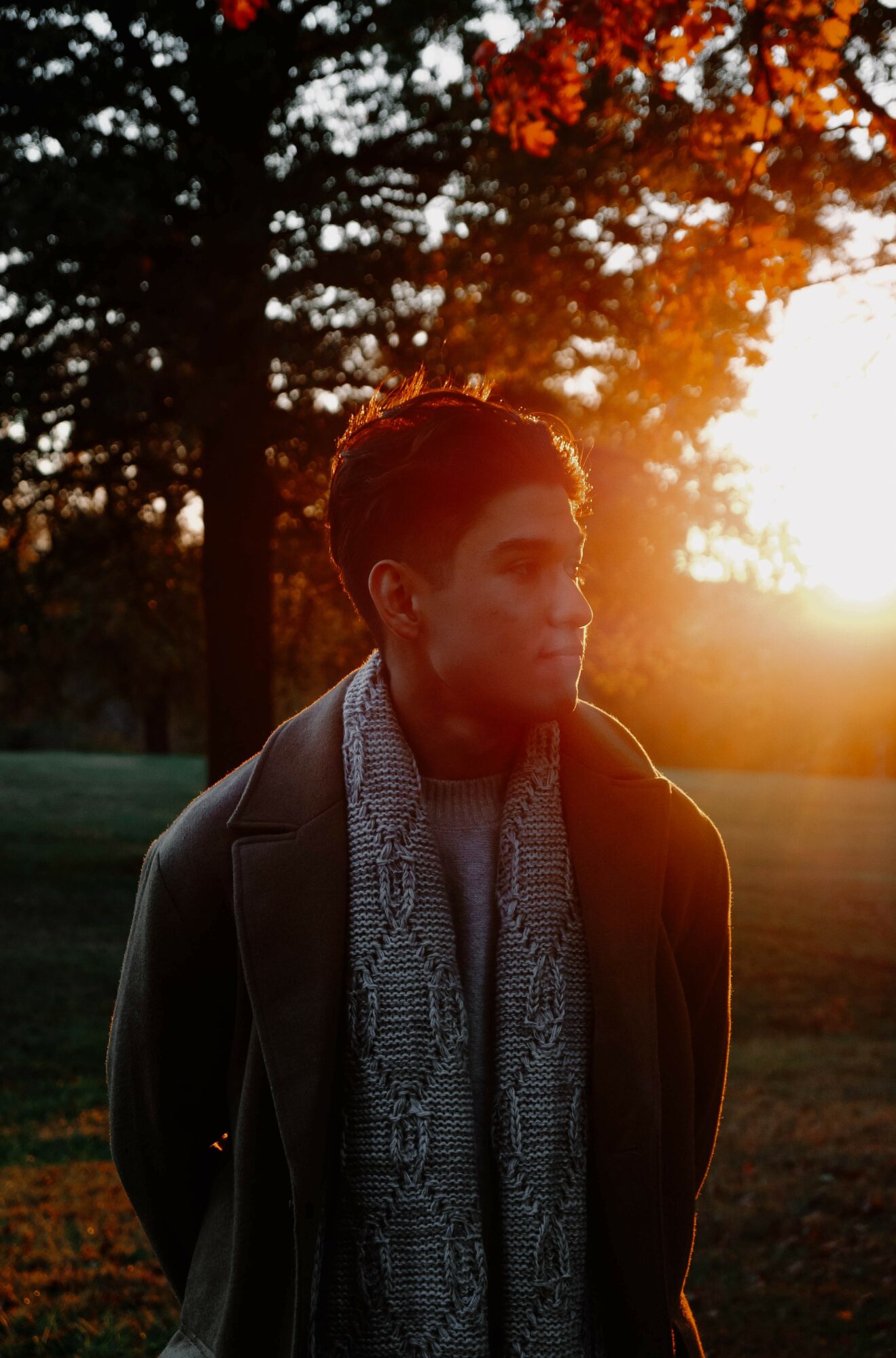 Woman standing outdoors during sunset with trees in background, wearing a coat and scarf, looking to the side.