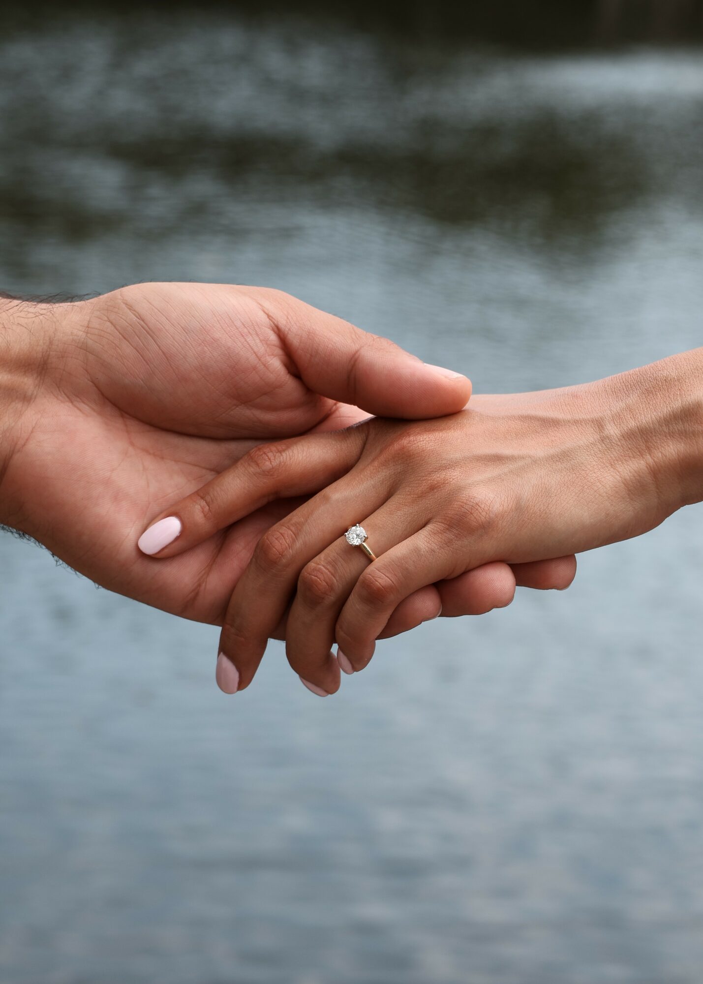 Two hands gently holding each other, one with a ring, against a blurred water background.