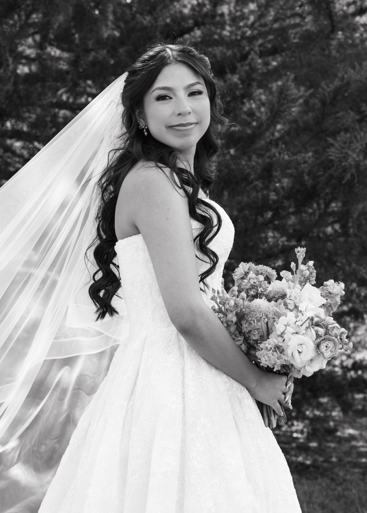 Bride in wedding dress holding bouquet outdoors, smiling, with trees in background.