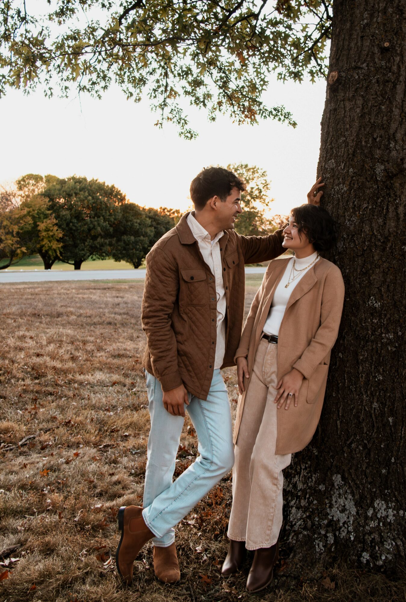 Two people leaning against a tree outdoors, looking at each other, with trees and open field in background.